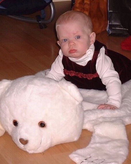 Six-month-old Tiegan-Elise crawling on a bear-shaped rug; she wears a white polo-necked jumper under a wine-coloured velvet dress.