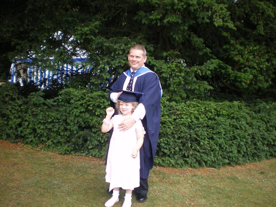 Six-Year-Old Tiegan-Elise on her dad’s graduation, behind her dad has one hand cuddling her and the other putting his graduation cap on her head.