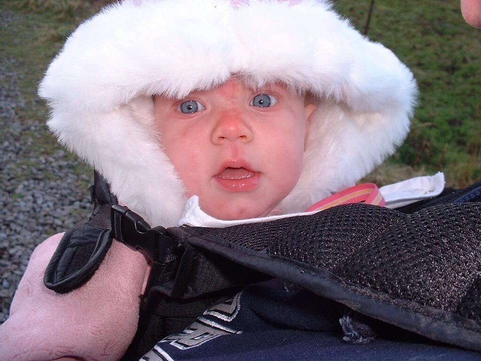 Three-month-old Tiegan-Elise wears a dusky-pink coat with a thick white fur hood, she is in a sling.