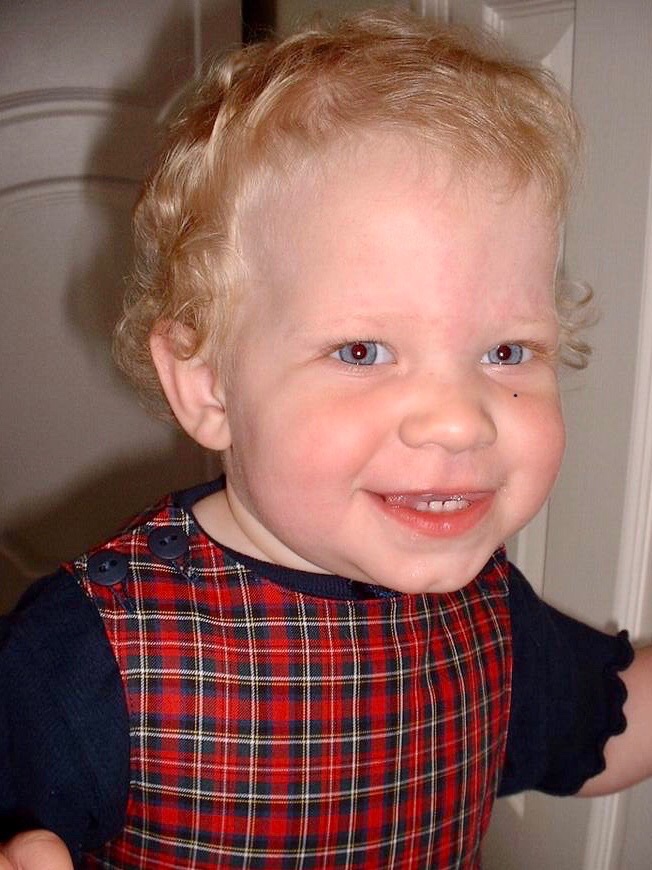 18-month-old Tiegan-Elise smiling, she wears a navy-blue t-shirt under a red, navy-blue and green checkered pinafore, she is in a bedroom.