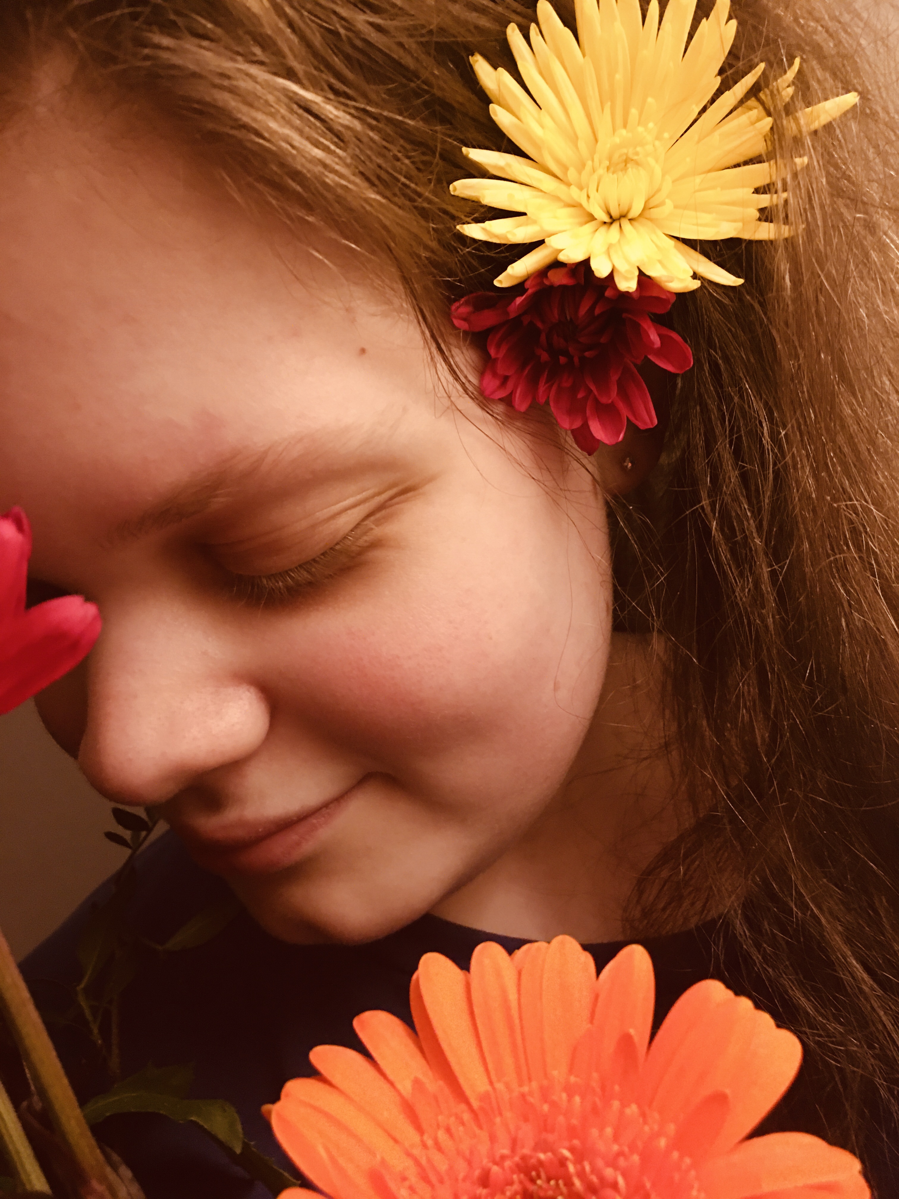 Fifteen Year Old Tiegan-Elise, she has flowers in her now-brunette wavy long hair and holds some other flowers in front of her face whilst looking down and smiling.
