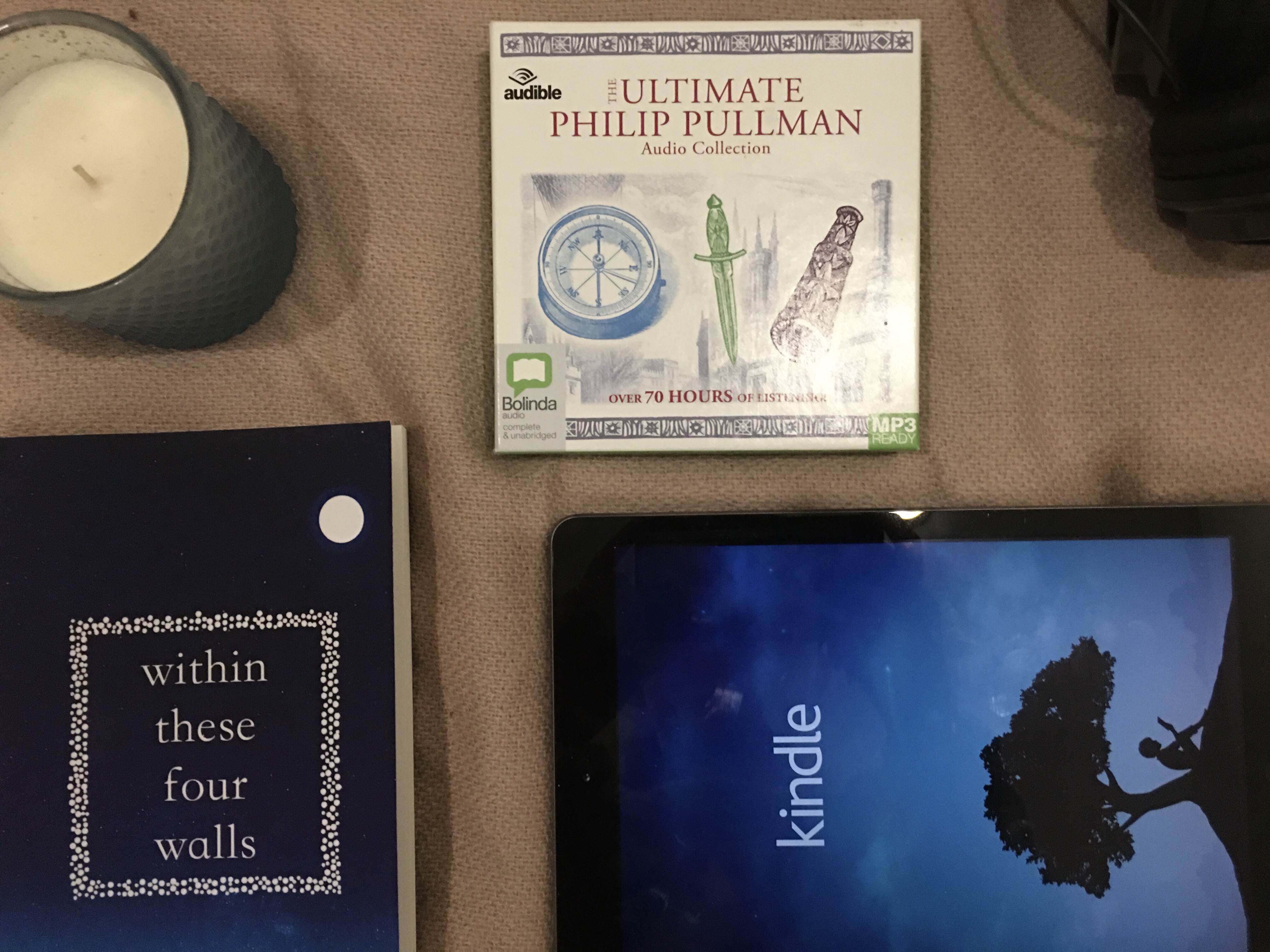 An audiobook set, a pair of headphones, an IPad displaying the kindle app, a paperback and a candle lay on a pale pink blanket 