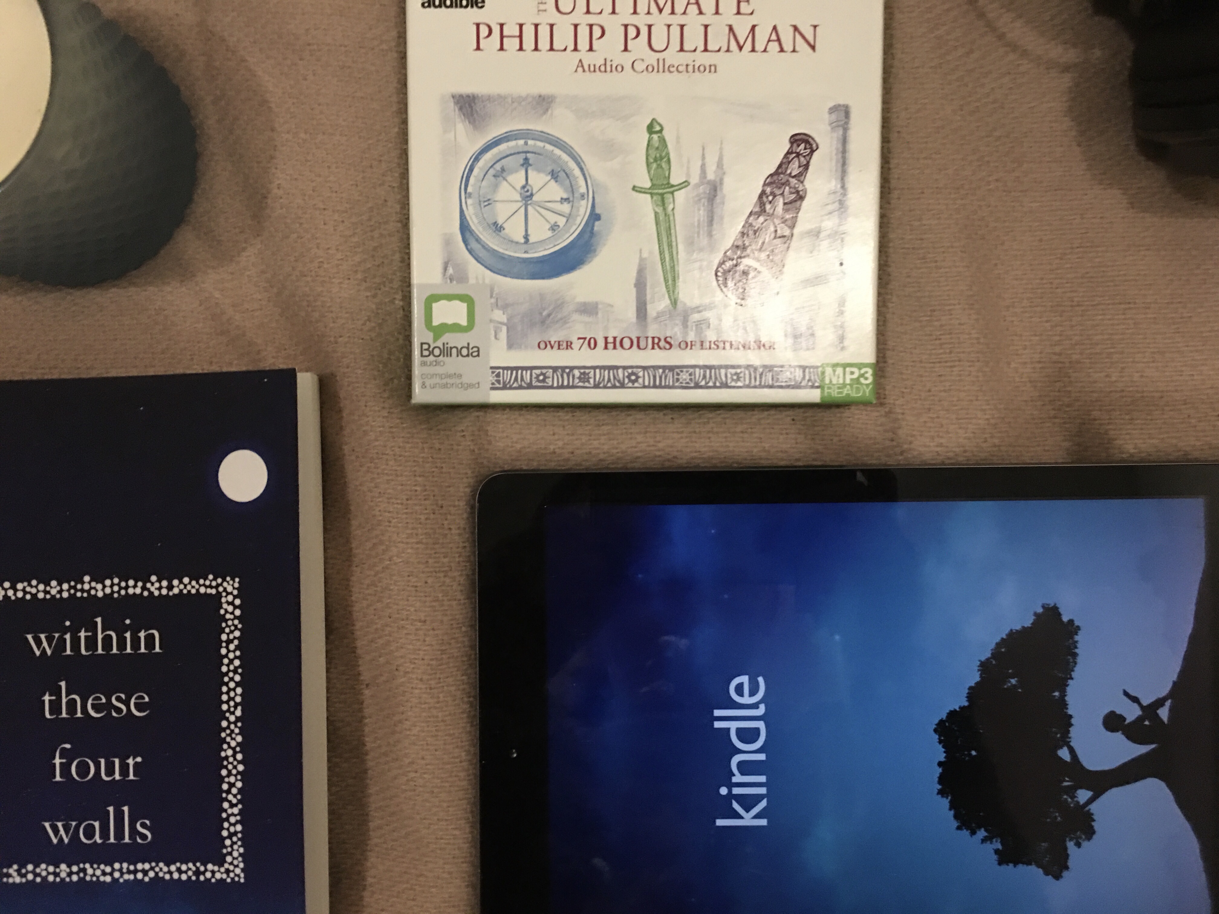 An audiobook set, a pair of headphones, an IPad displaying the kindle app, a paperback and a candle lay on a pale pink blanket 