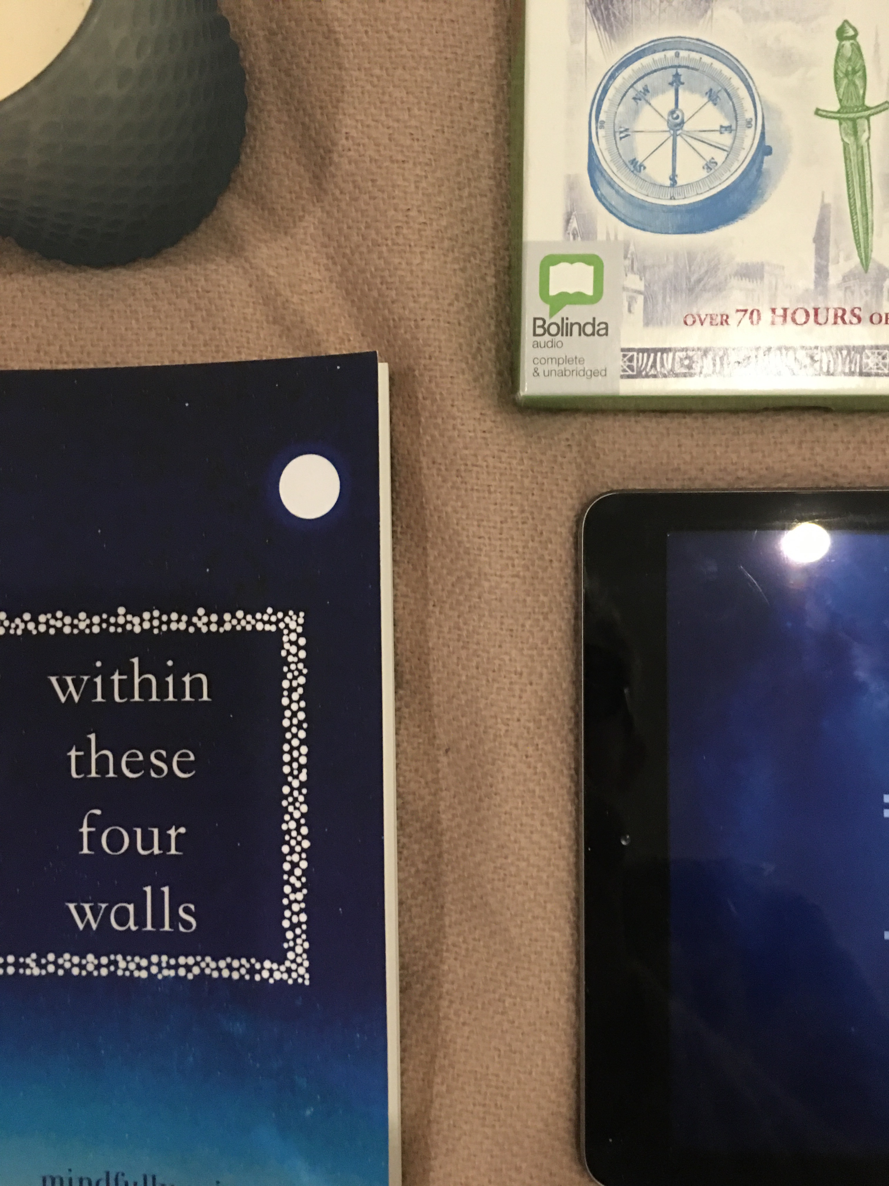 An audiobook set, a pair of headphones, an IPad displaying the kindle app, a paperback and a candle lay on a pale pink blanket 