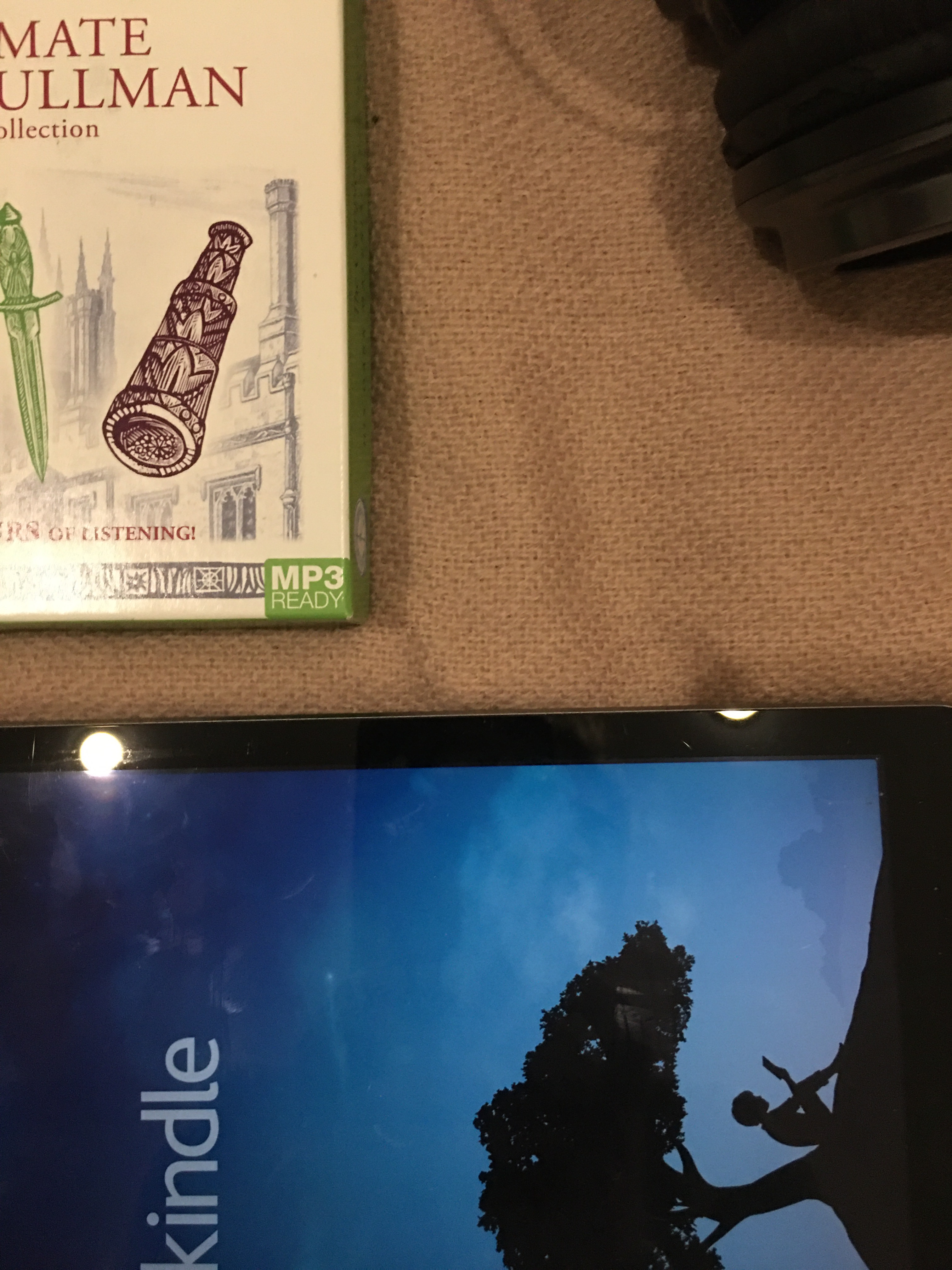 An audiobook set, an IPad displaying the kindle app, a paperback and a candle lay on a pale pink blanket 