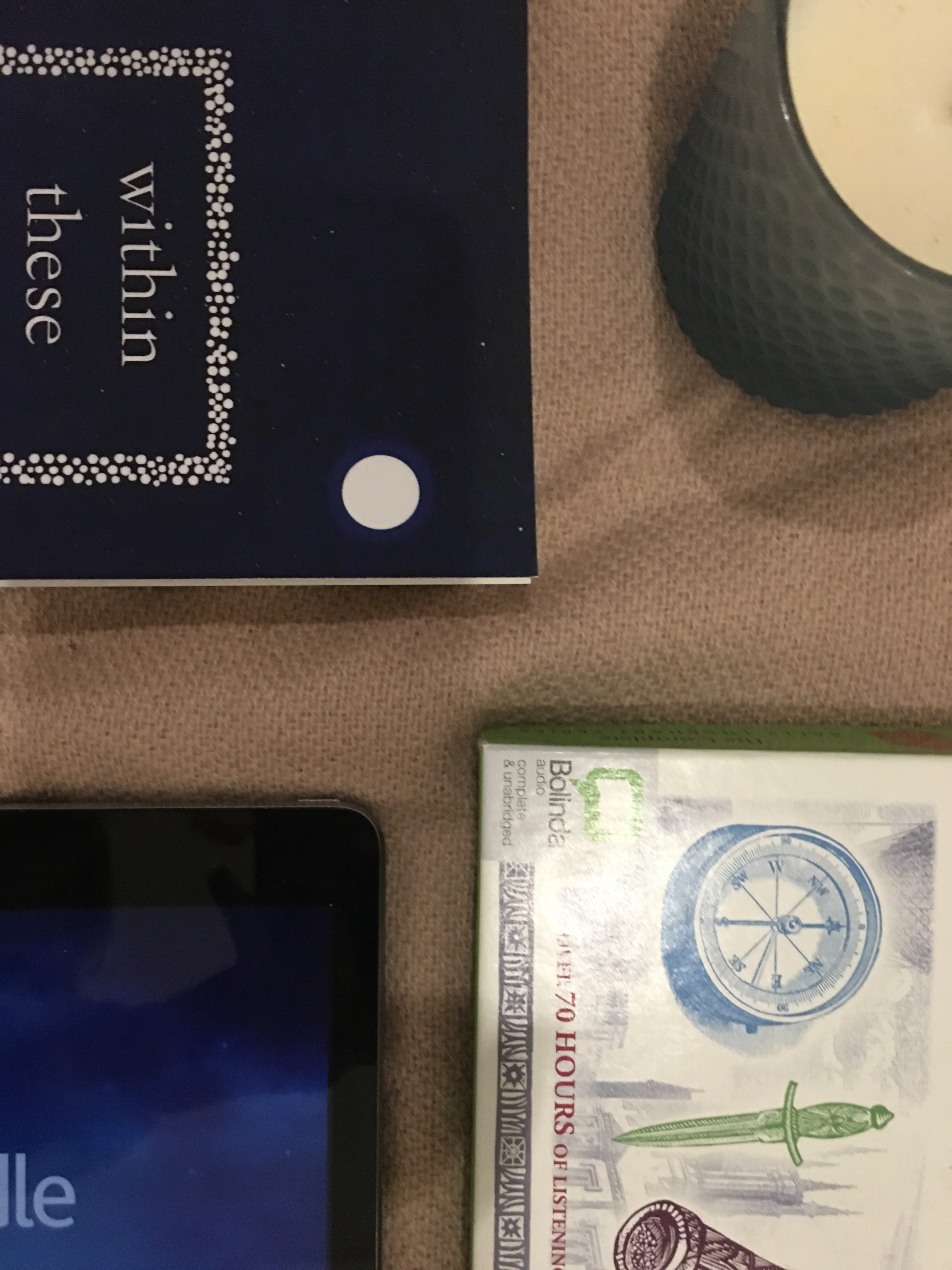 An audiobook set, a pair of headphones, an IPad displaying the kindle app, a paperback and a candle lay on a pale pink blanket 