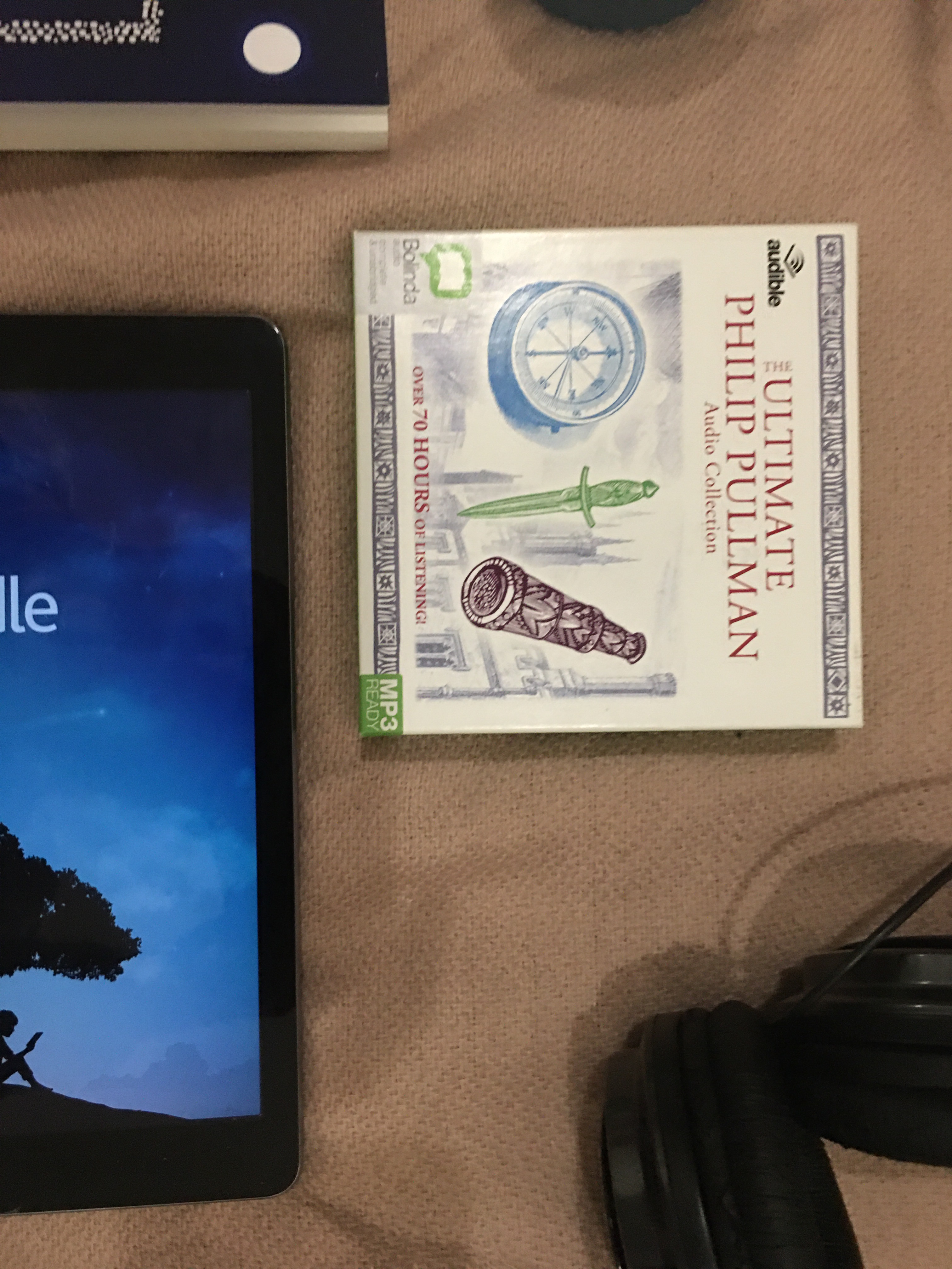 An audiobook set, a pair of headphones, an IPad displaying the kindle app, a paperback and a candle lay on a pale pink blanket 