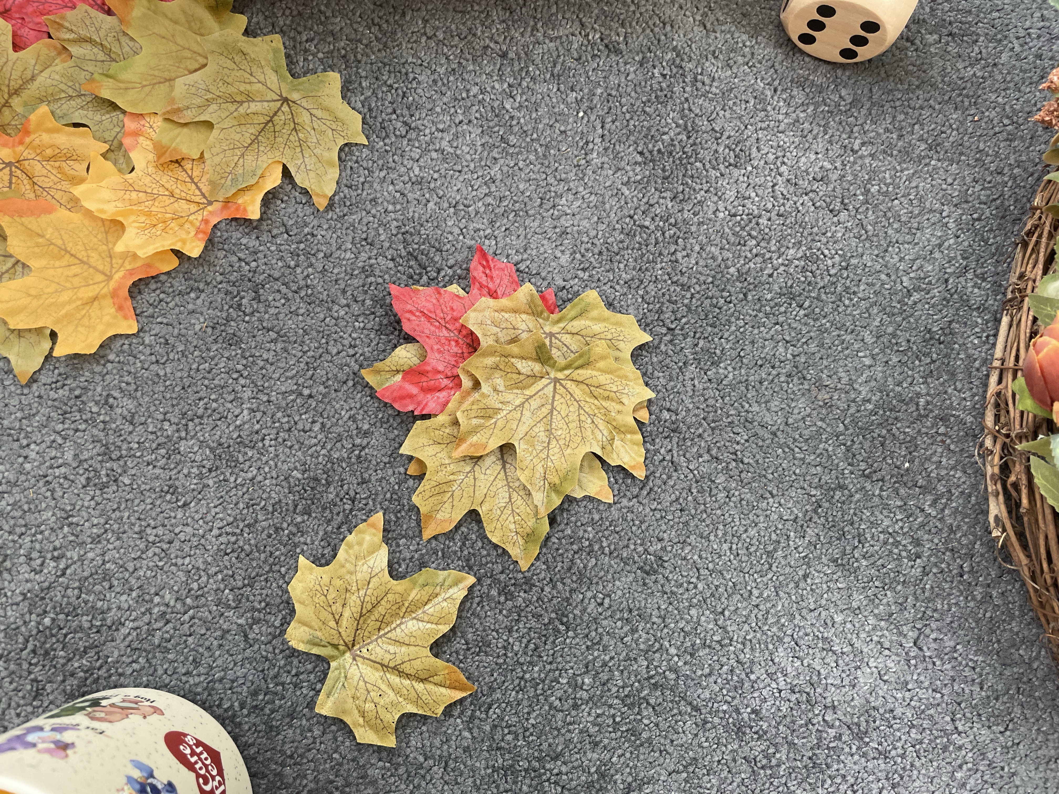 Some faux leaves lay by a wooden dice, a self care bear mug and an faux Autumnal coloured floral wreath on a charcoal carpet.