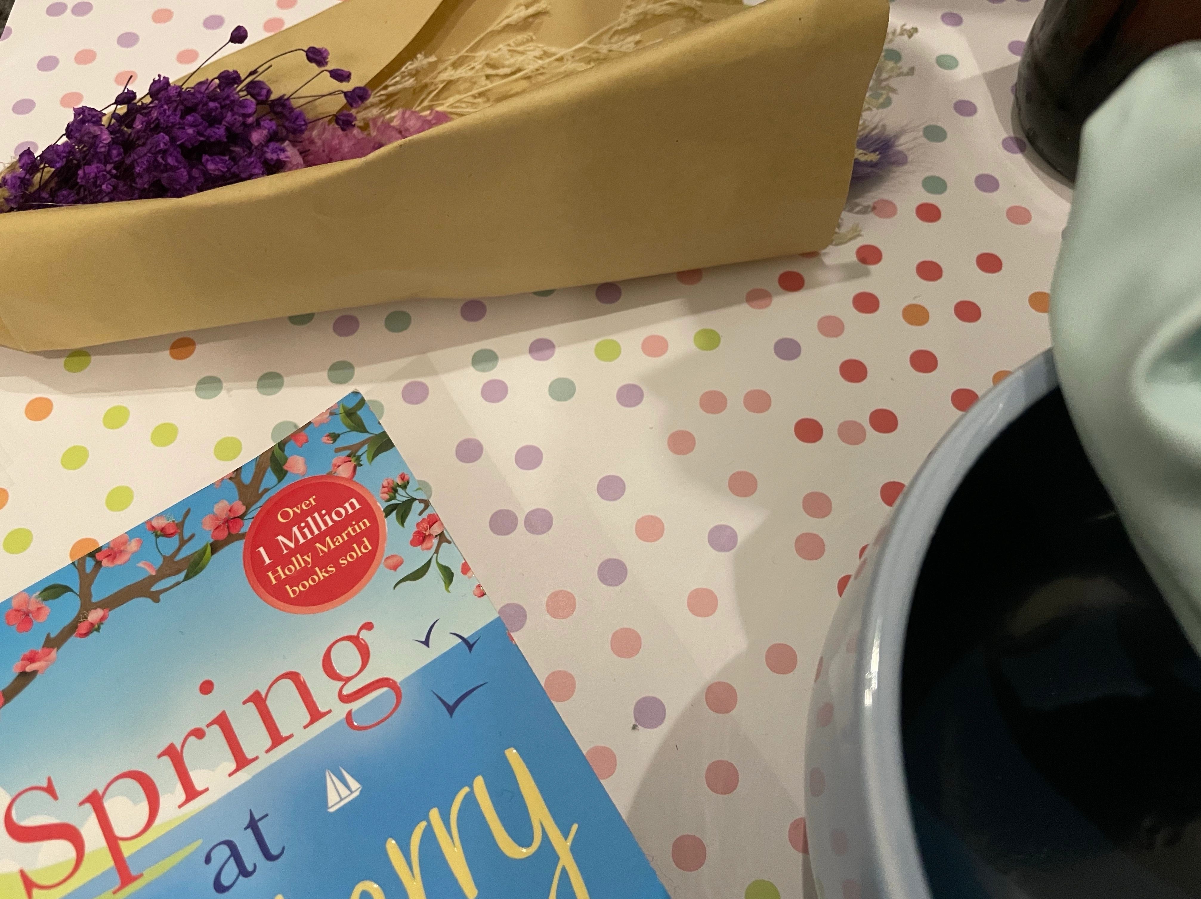 A bouquet of purple dried flowers lay beside items such as a two-toned blue smiley face yarn bowl, a baby blue faux leather scrunchie and the Spring At Blueberry Bay paperback on a pastel multicoloured polka dot background.