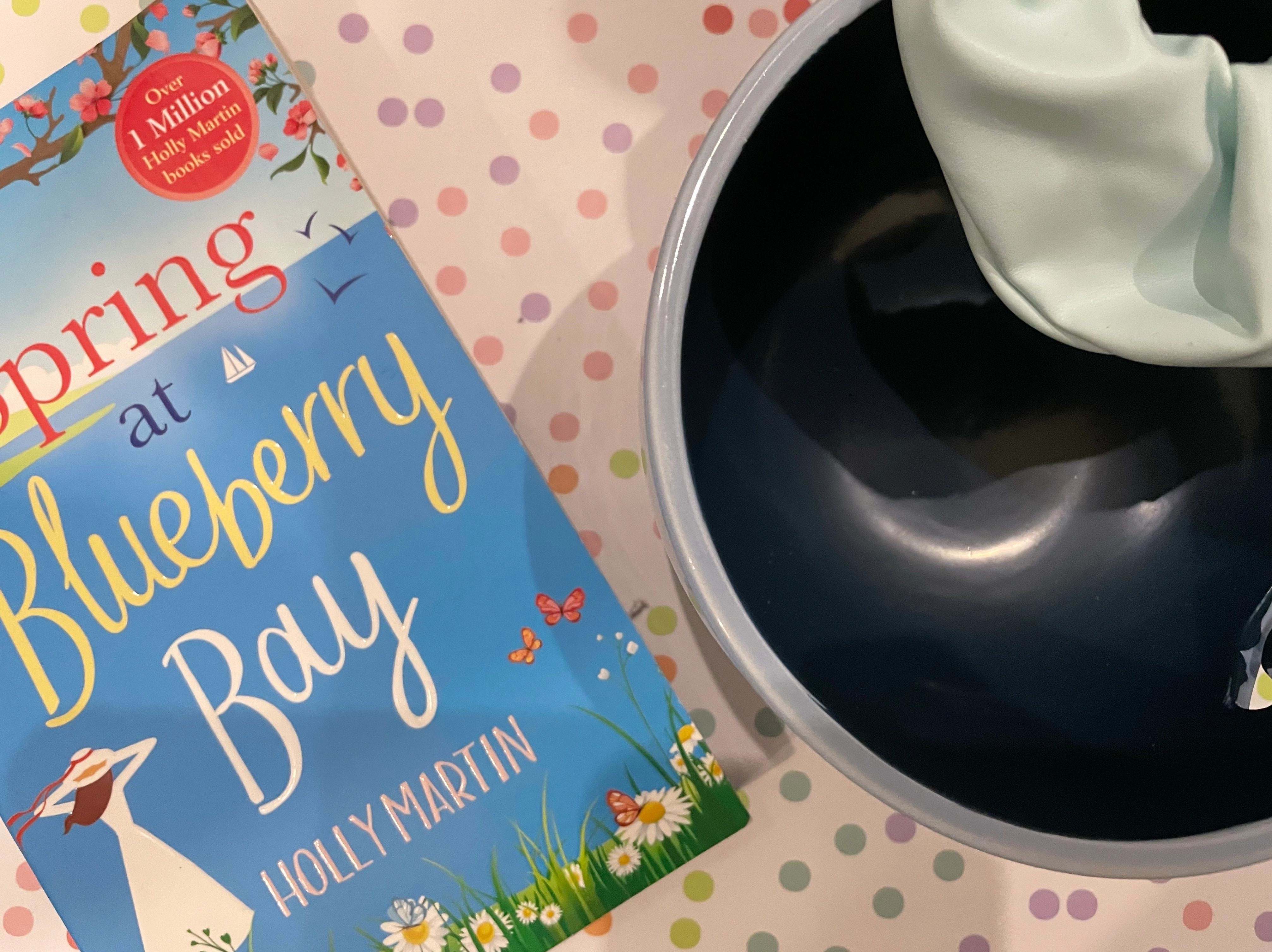 A bouquet of purple dried flowers lay beside items such as a two-toned blue smiley face yarn bowl, a baby blue faux leather scrunchie and the Spring At Blueberry Bay paperback on a pastel multicoloured polka dot background.