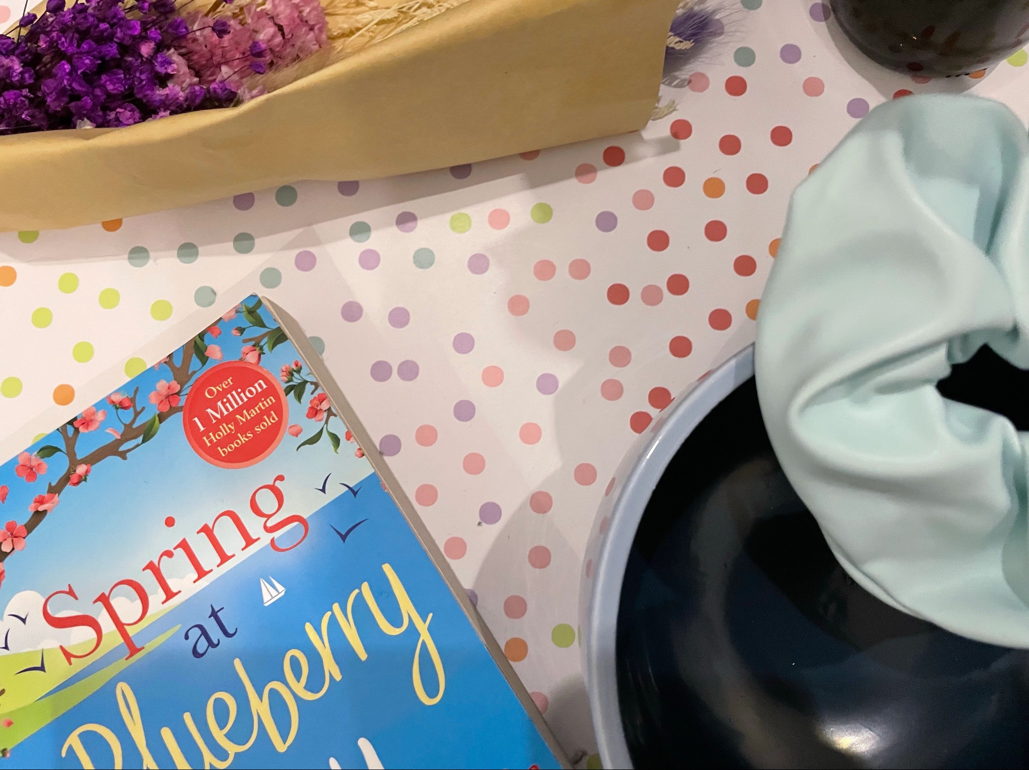 A bouquet of purple dried flowers lay beside items such as a two-toned blue smiley face yarn bowl, a baby blue faux leather scrunchie and the Spring At Blueberry Bay paperback on a pastel multicoloured polka dot background.