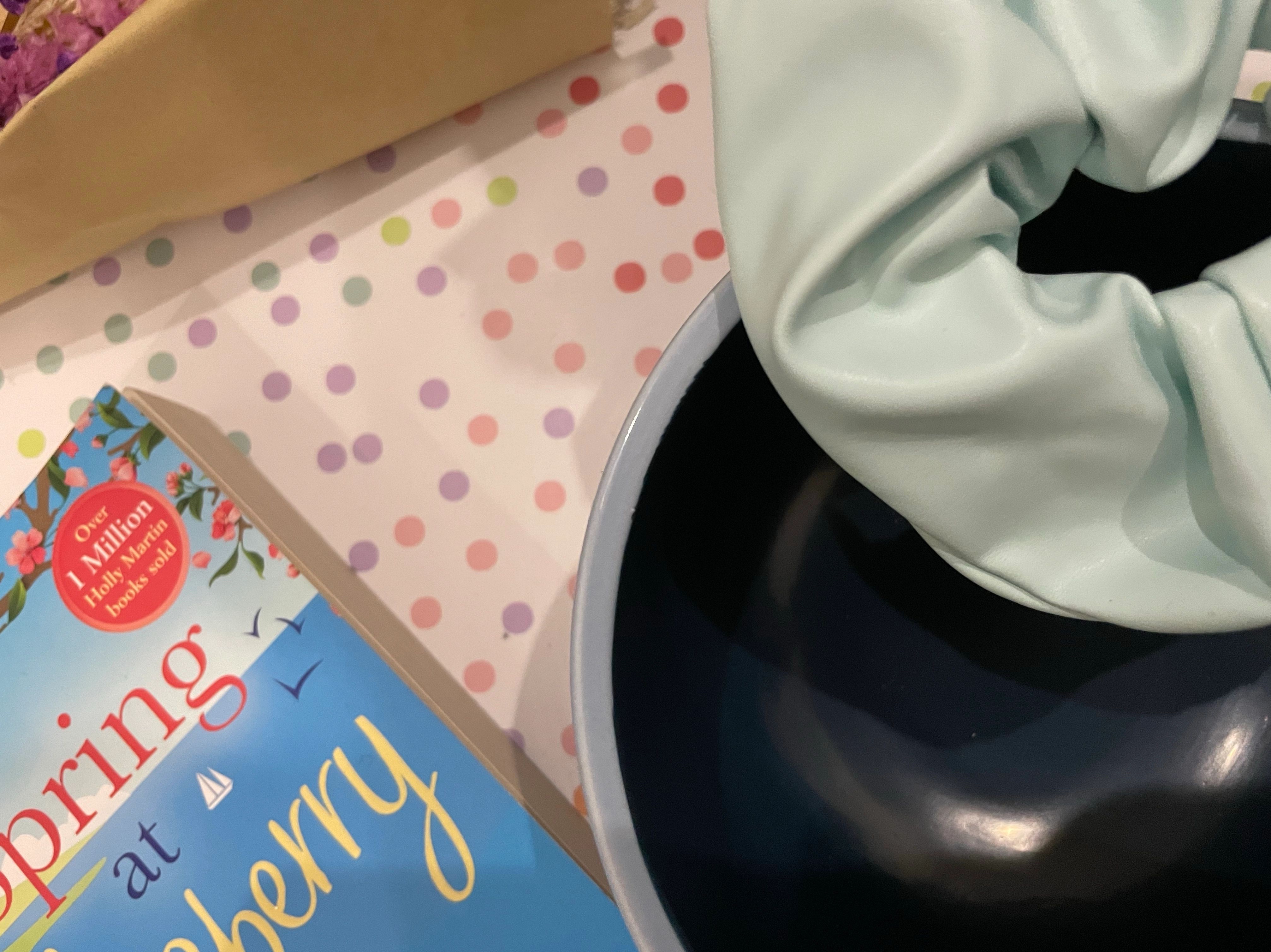 A bouquet of purple dried flowers lay beside items such as a two-toned blue smiley face yarn bowl, a baby blue faux leather scrunchie and the Spring At Blueberry Bay paperback on a pastel multicoloured polka dot background.