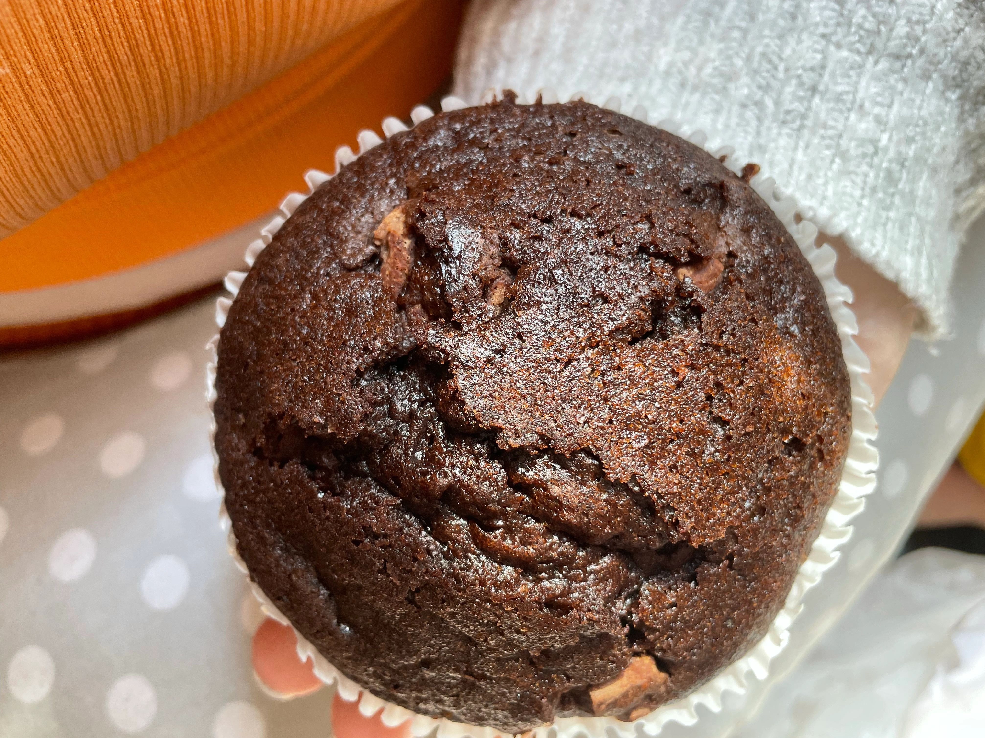 Tig’s hand holds a giant muffin upon their u-cushion that sits around them and has a grey and white polka dot cover. Their grey cardigan and orange cropped top are visible. 