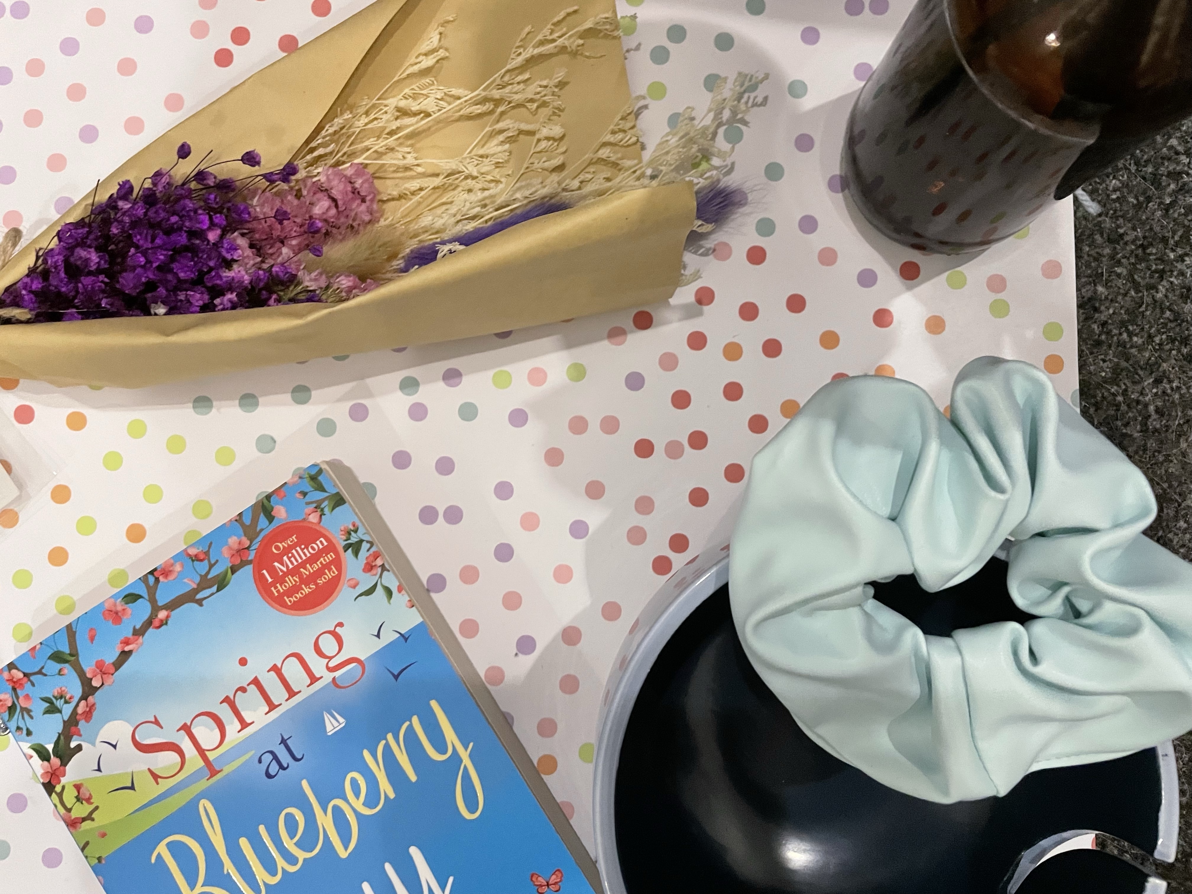 A bouquet of purple dried flowers lay beside items such as a two-toned blue smiley face yarn bowl, a baby blue faux leather scrunchie and the Spring At Blueberry Bay paperback on a pastel multicoloured polka dot background.