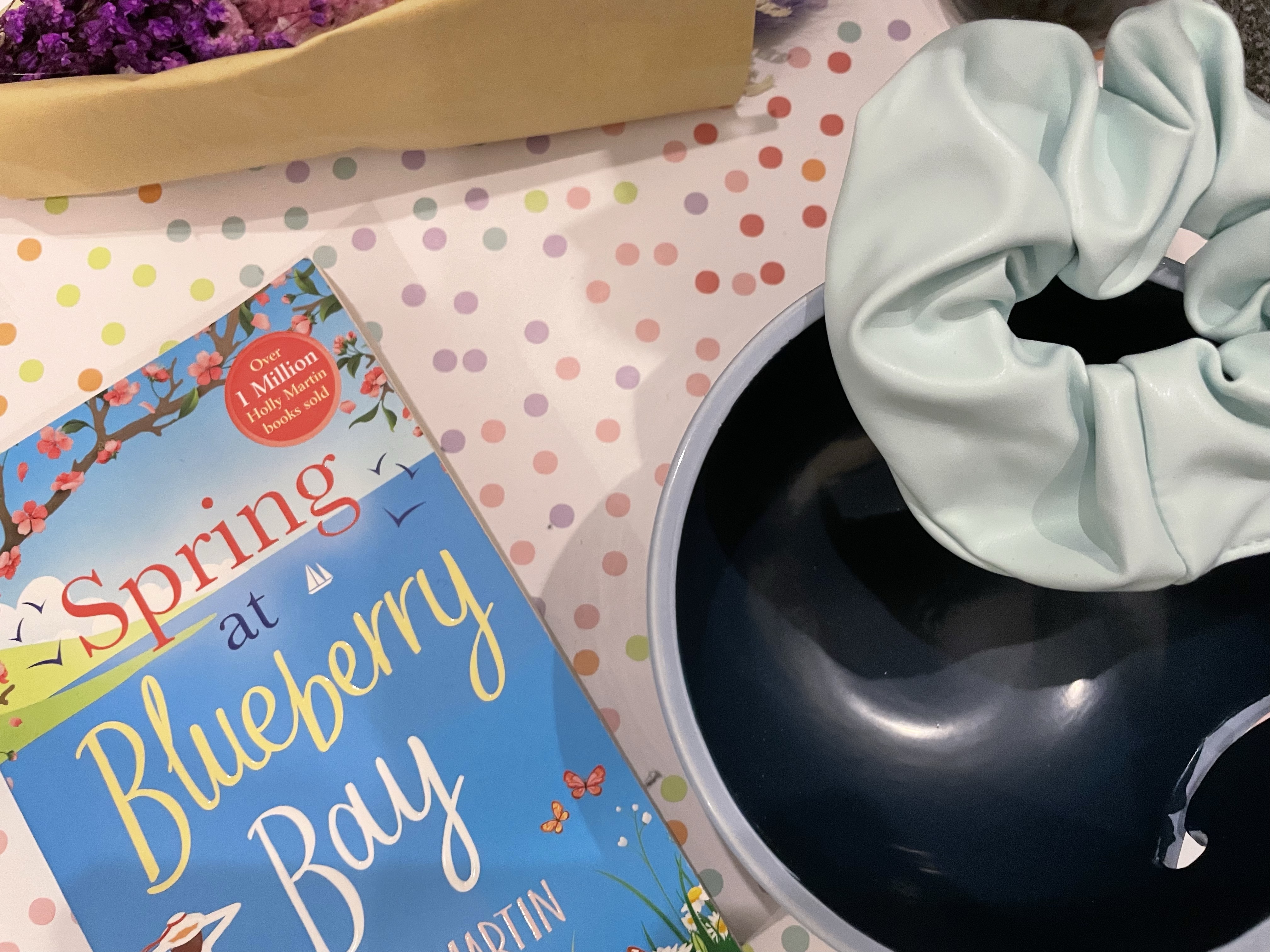 A bouquet of purple dried flowers lay beside items such as a two-toned blue smiley face yarn bowl, a baby blue faux leather scrunchie and the Spring At Blueberry Bay paperback on a pastel multicoloured polka dot background.