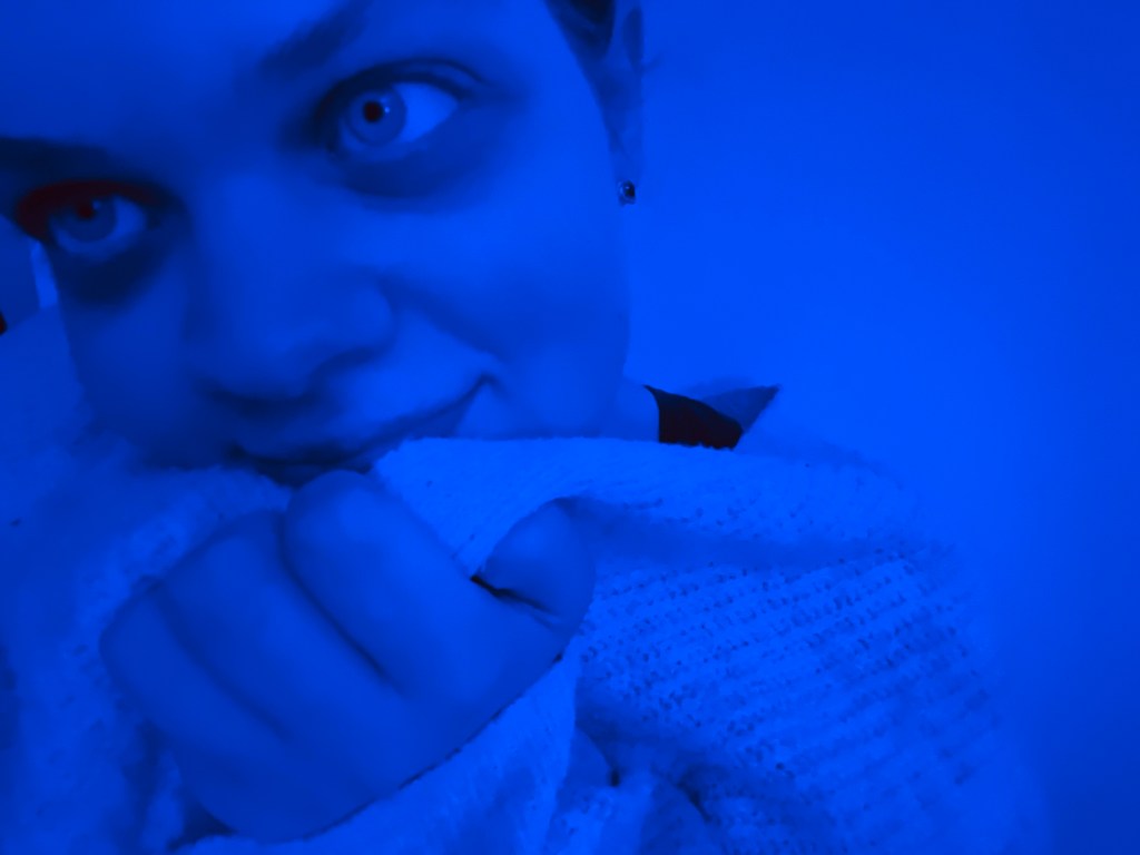 Tig cuddles a cardigan into their face whilst sat in a blue light.