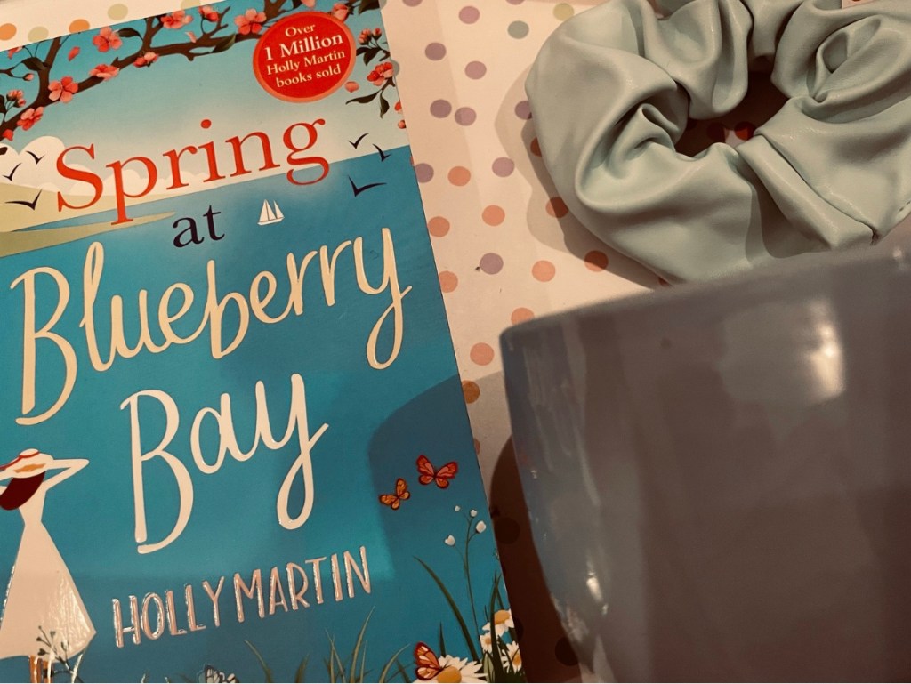 a bouquet of dried purple dyed flowers lays beside a two-toned yarn bowl, a baby blue scrunchie and other items on a pastel polka dot background.