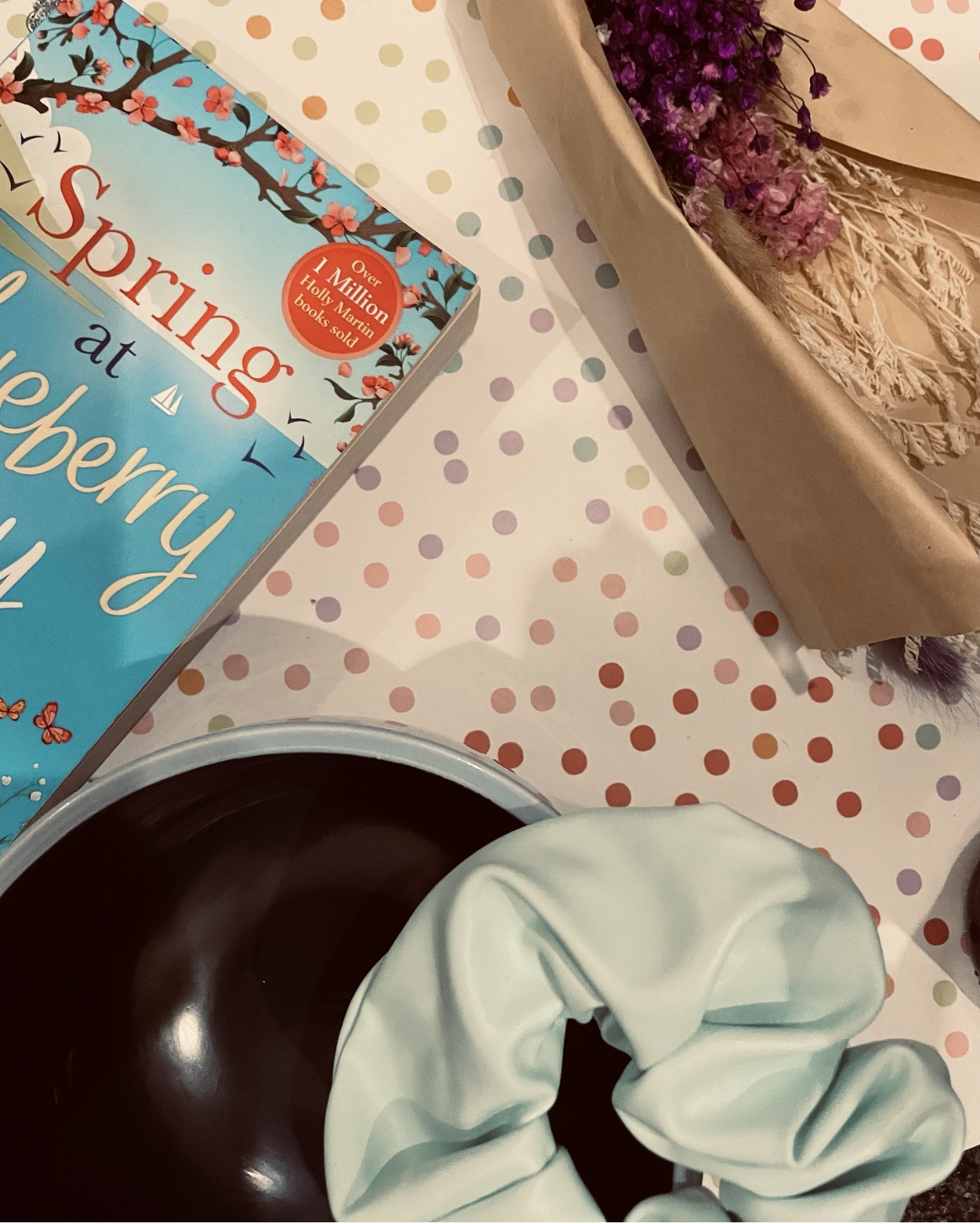 a bouquet of dried purple dyed flowers lays beside a two-toned yarn bowl, a baby blue scrunchie and other items on a pastel polka dot background.