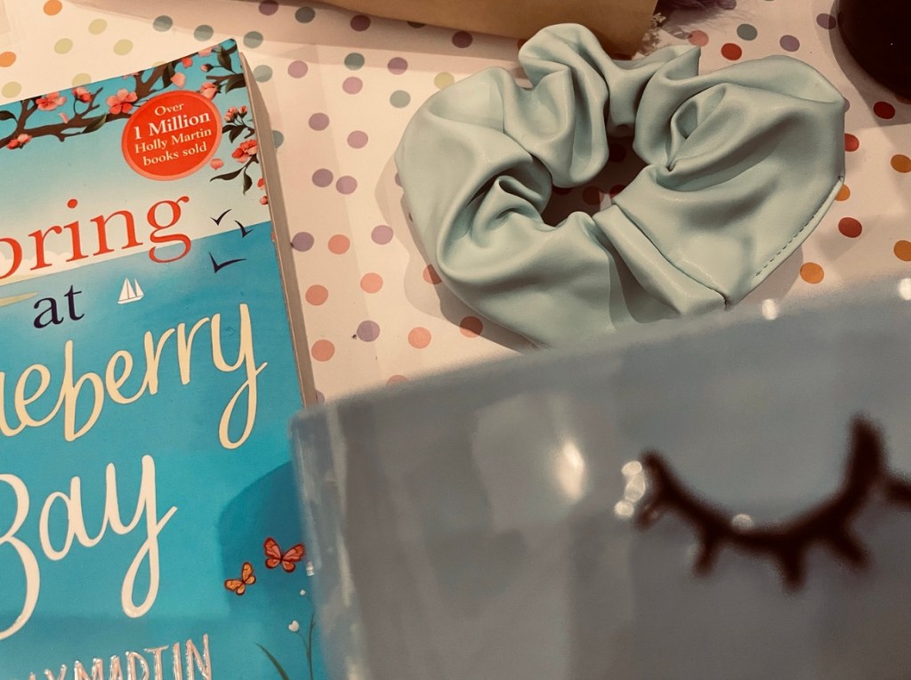 a bouquet of dried purple dyed flowers lays beside a two-toned yarn bowl, a baby blue scrunchie and other items on a pastel polka dot background.