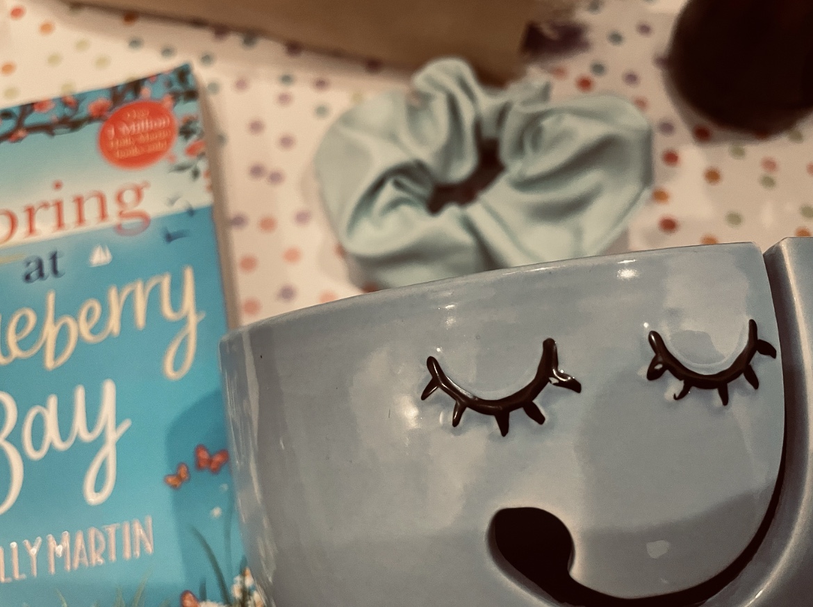 A two-toned blue smiley face yarn bowl beside items such as a bouquet of purple dried flowers lay, a baby blue faux leather scrunchie and the Spring At Blueberry Bay paperback on a pastel multicoloured polka dot background.