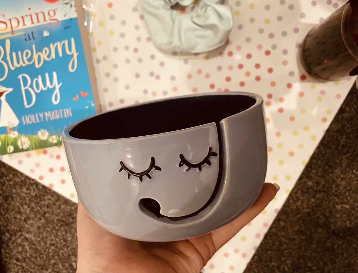  A blue smiley face yarn bowl held above items including the Spring At Blueberry Bay book on a pastel polka dot background.