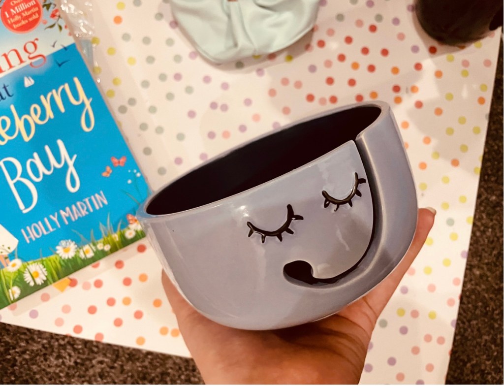 a bouquet of dried purple dyed flowers lays beside a two-toned yarn bowl, a baby blue scrunchie and other items on a pastel polka dot background.