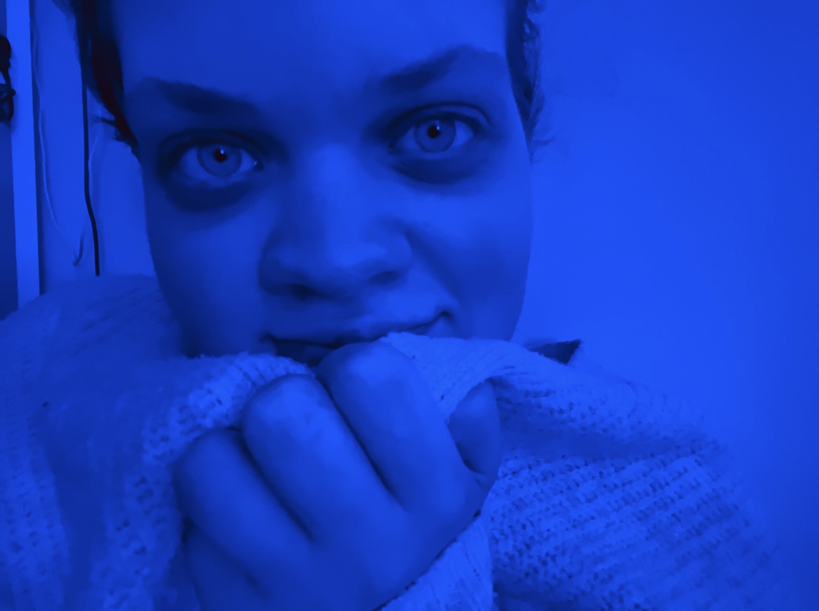 Tig cuddles a cardigan into their face whilst sat in a blue light.