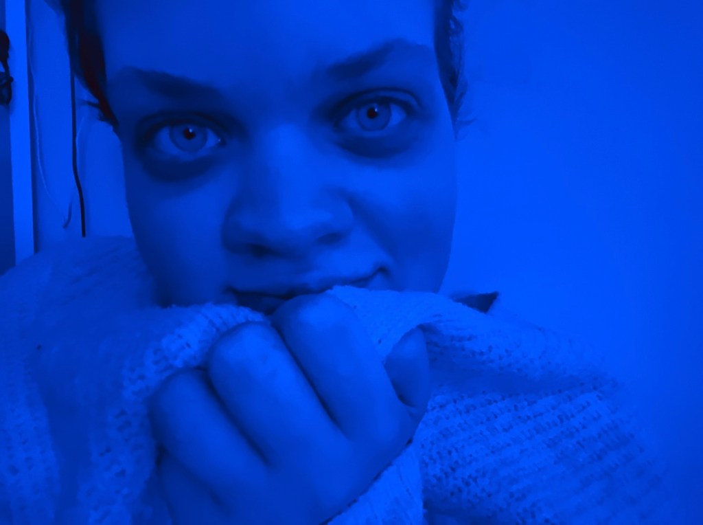 Tig cuddles a cardigan into their face whilst sat in a blue light.