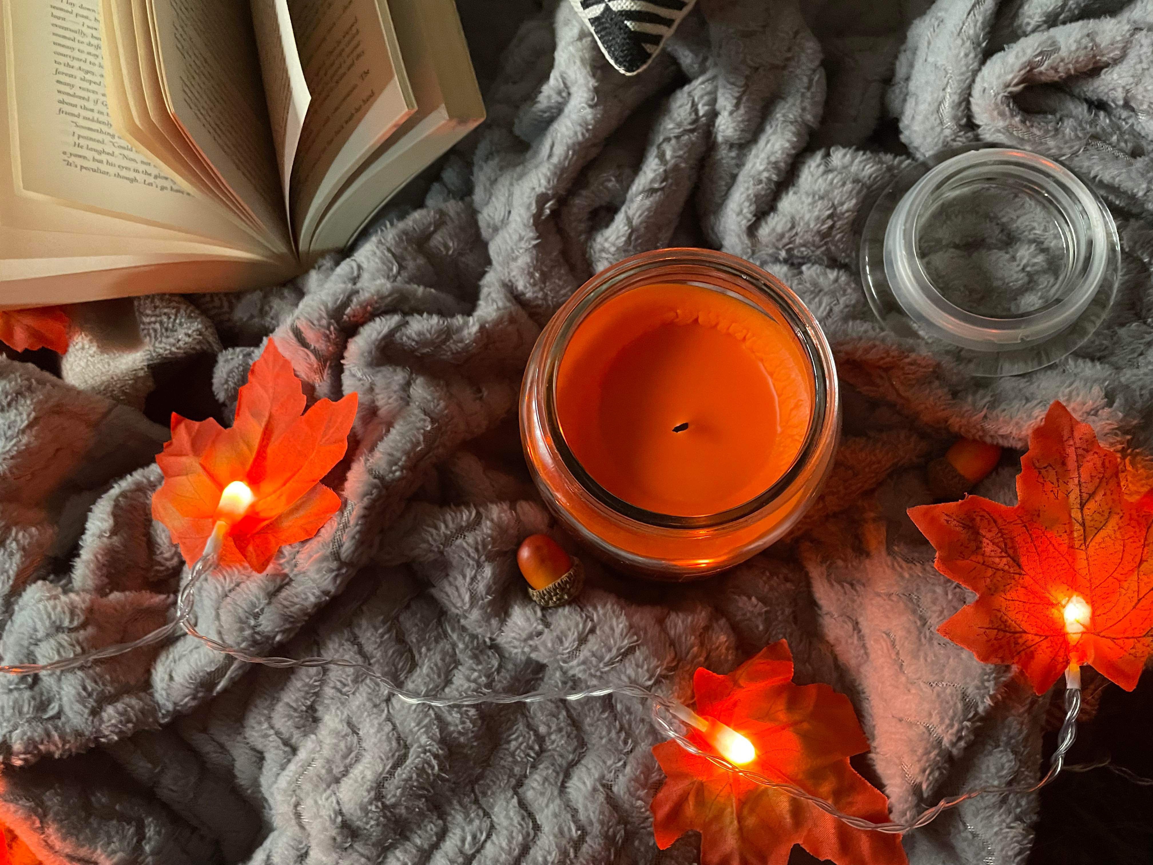 A candle, book, Autumn leaf LED lights and other autumnal items on a grey blanket that lays on a tartan throw.