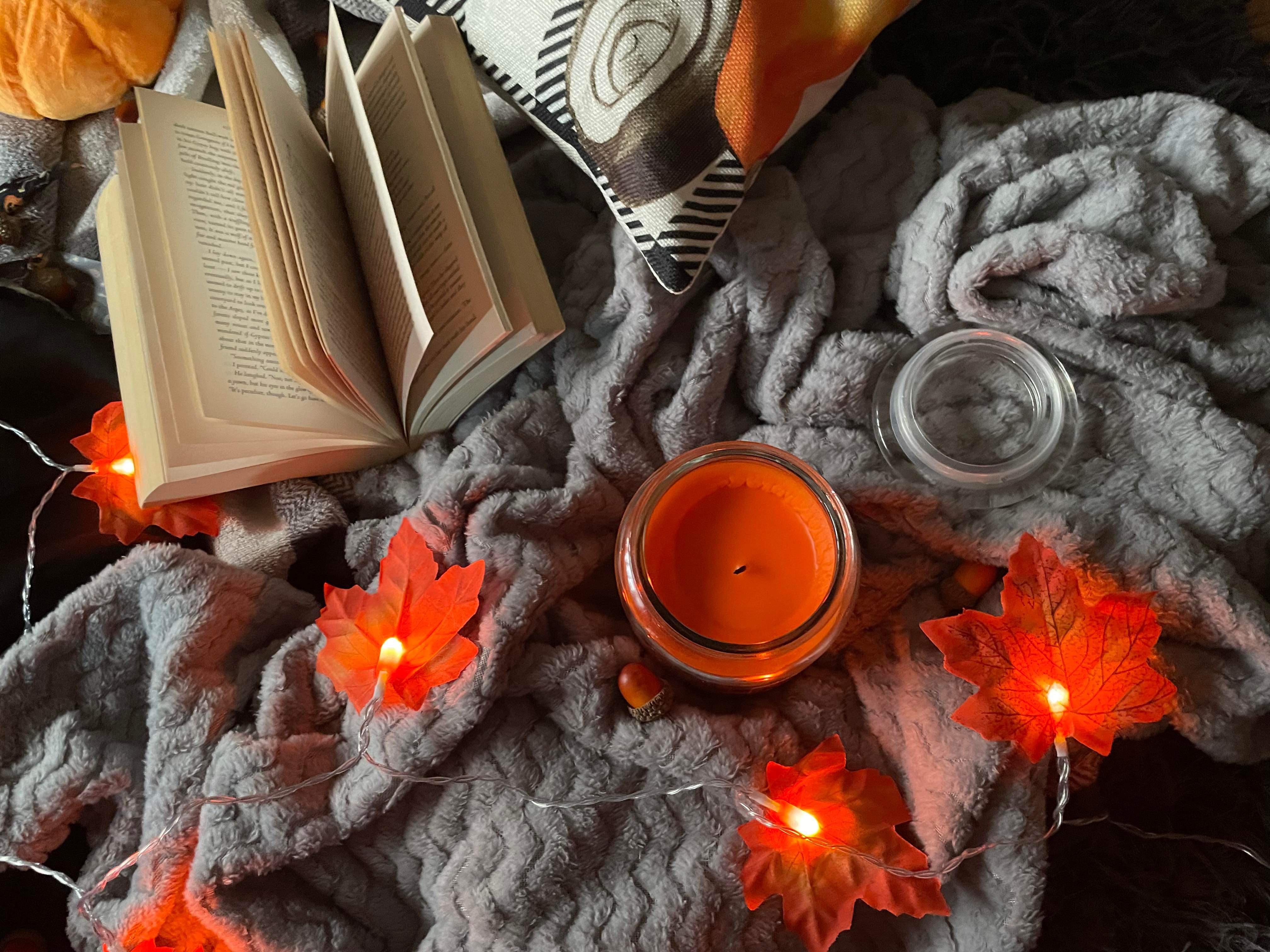 A candle, book, Autumn leaf LED lights and other autumnal items on a grey blanket that lays on a tartan throw.