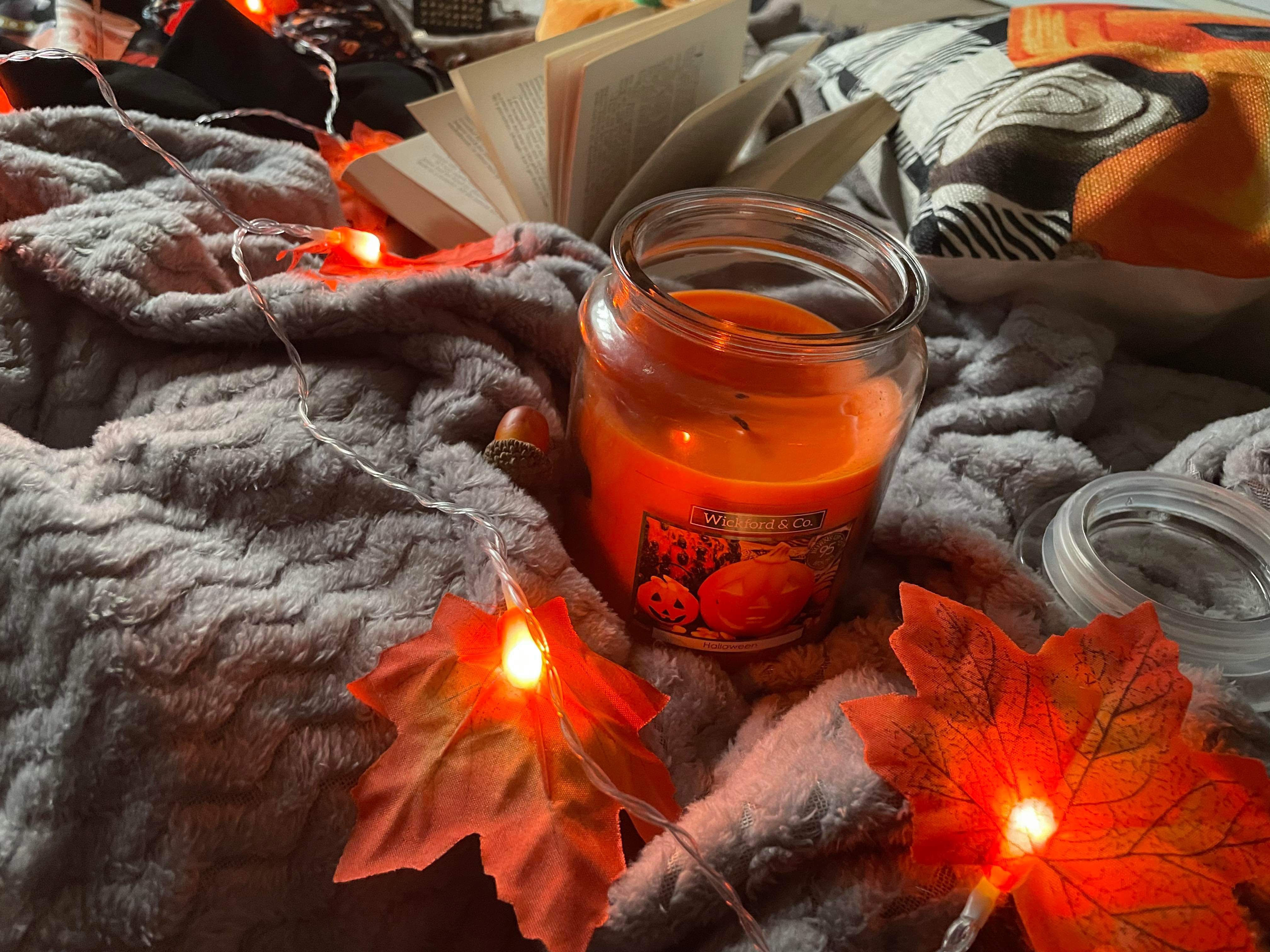 A candle, book, Autumn leaf LED lights and other autumnal items on a grey blanket that lays on a tartan throw.