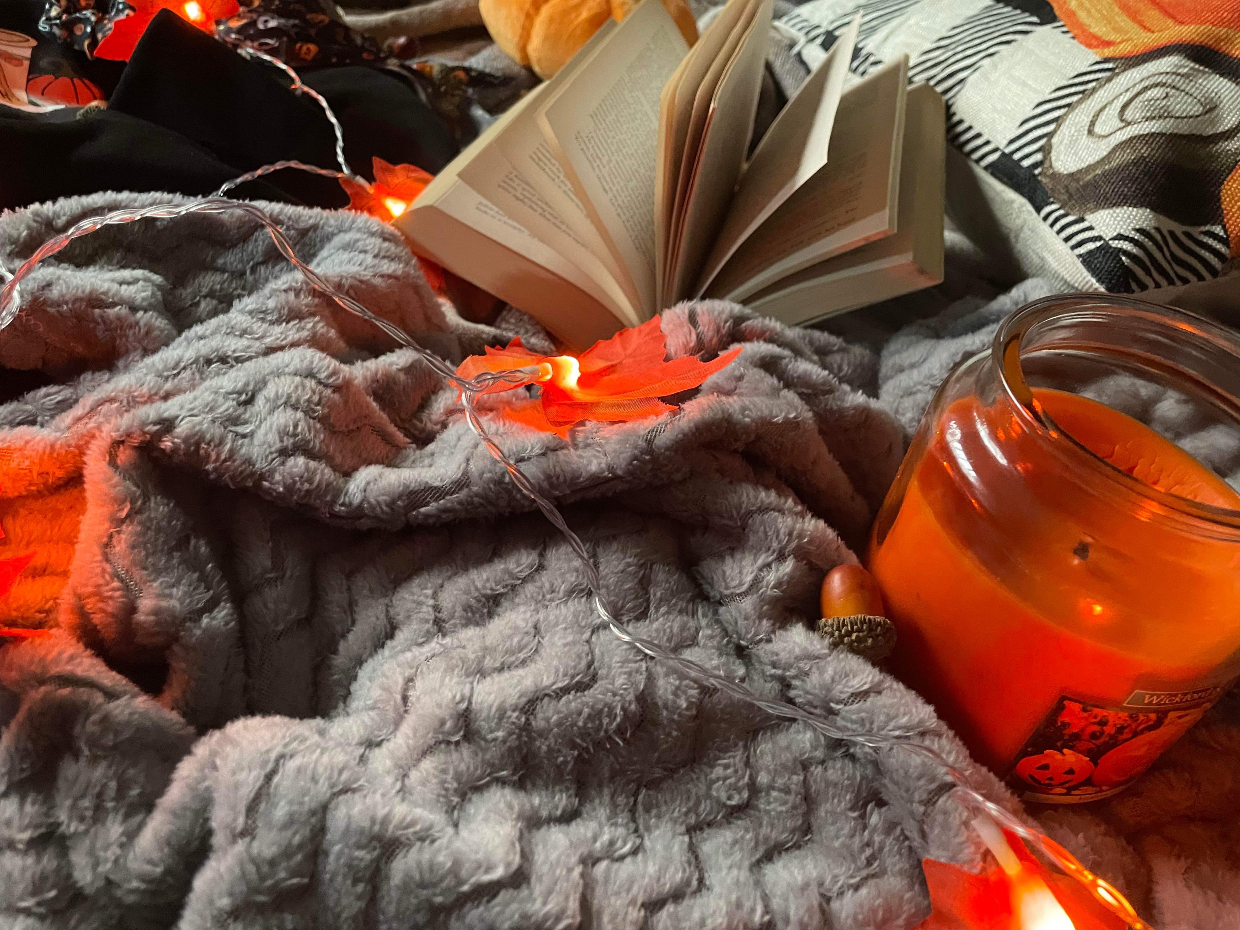 A candle, book, Autumn leaf LED lights and other autumnal items on a grey blanket that lays on a tartan throw.