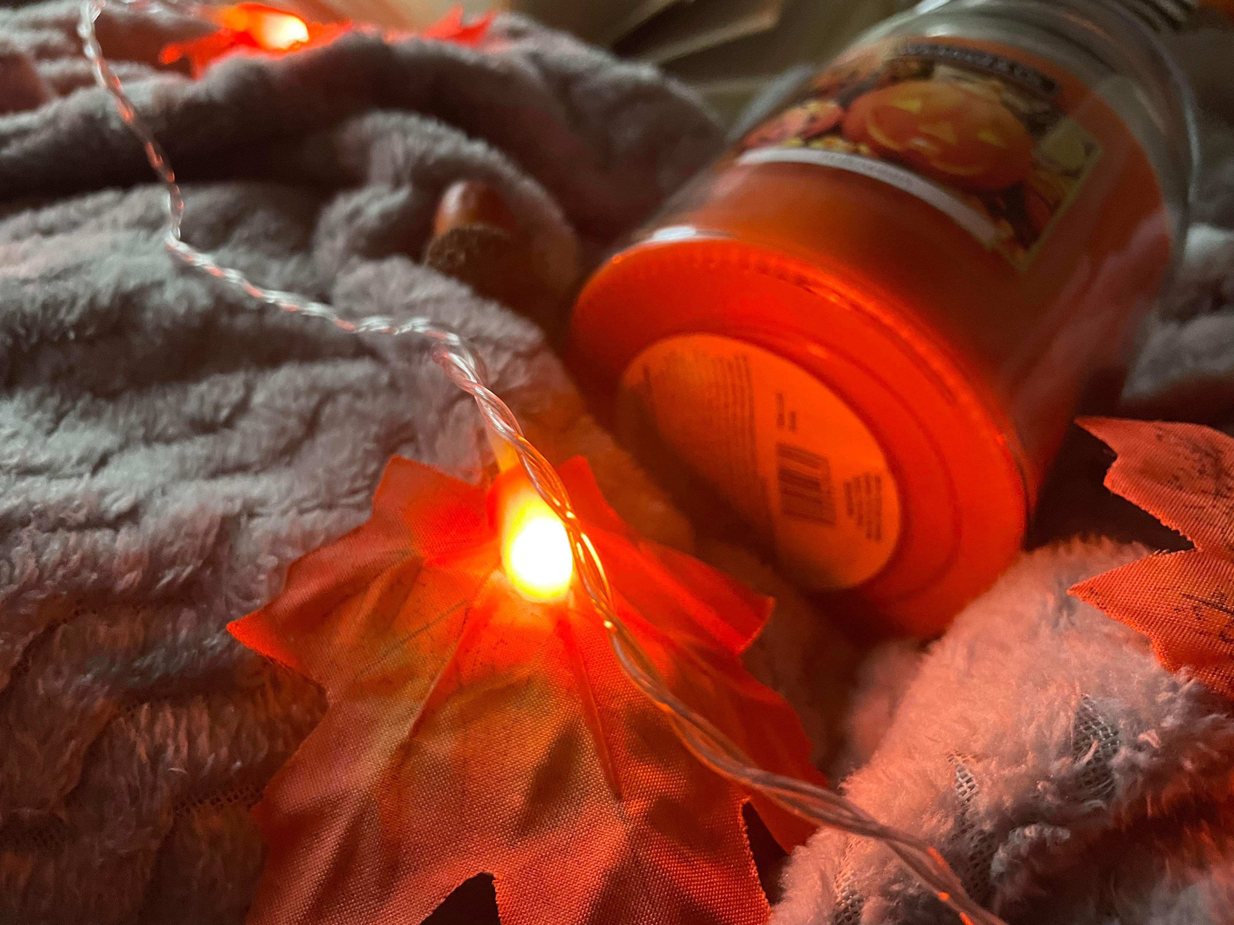 A Halloween candle, LED faux autumn leaf lights and other autumnal items on a grey fleece blanket 