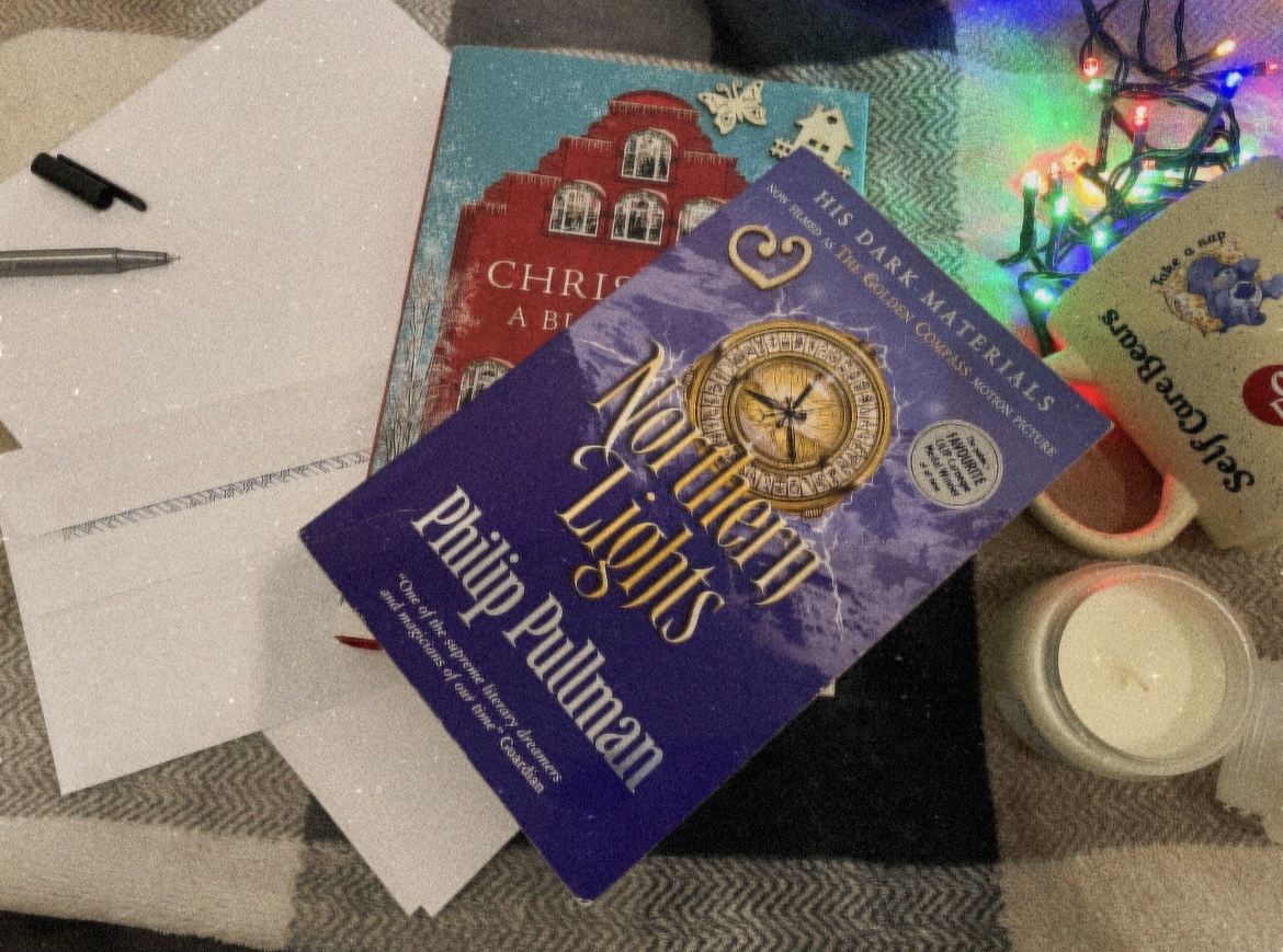 Fairy lights in a mug, letter writing material, a candle, a Christmas book and Philip Pullman’s “Northern Lights” on a tartan throw