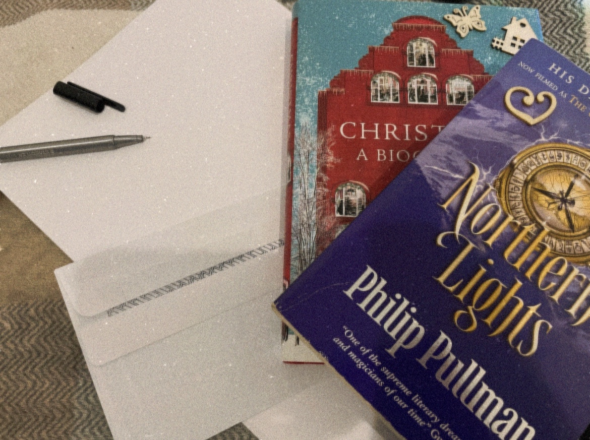 Fairy lights in a mug, letter writing material, a candle, a Christmas book and Philip Pullman’s “Northern Lights” on a tartan throw