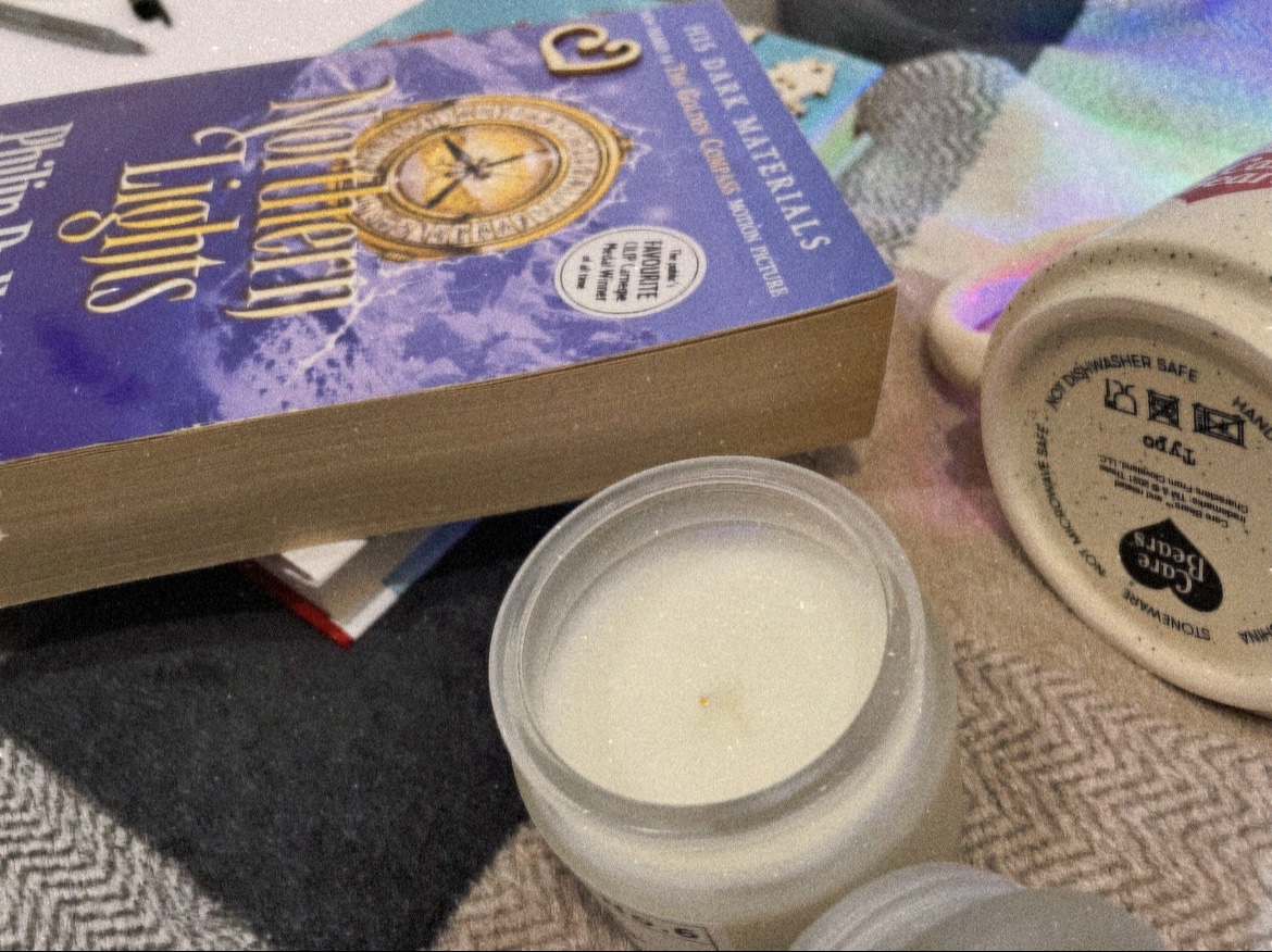 Fairy lights in a mug, letter writing material, a candle, a Christmas book and Philip Pullman’s “Northern Lights” on a tartan throw