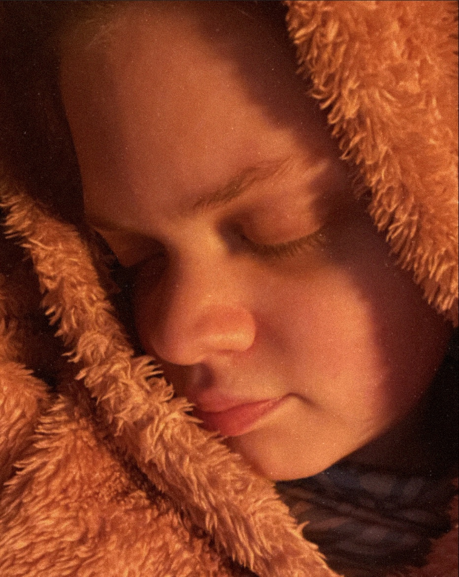 Tig in a pink fur hoodie faces away from the camera with their eyes shut