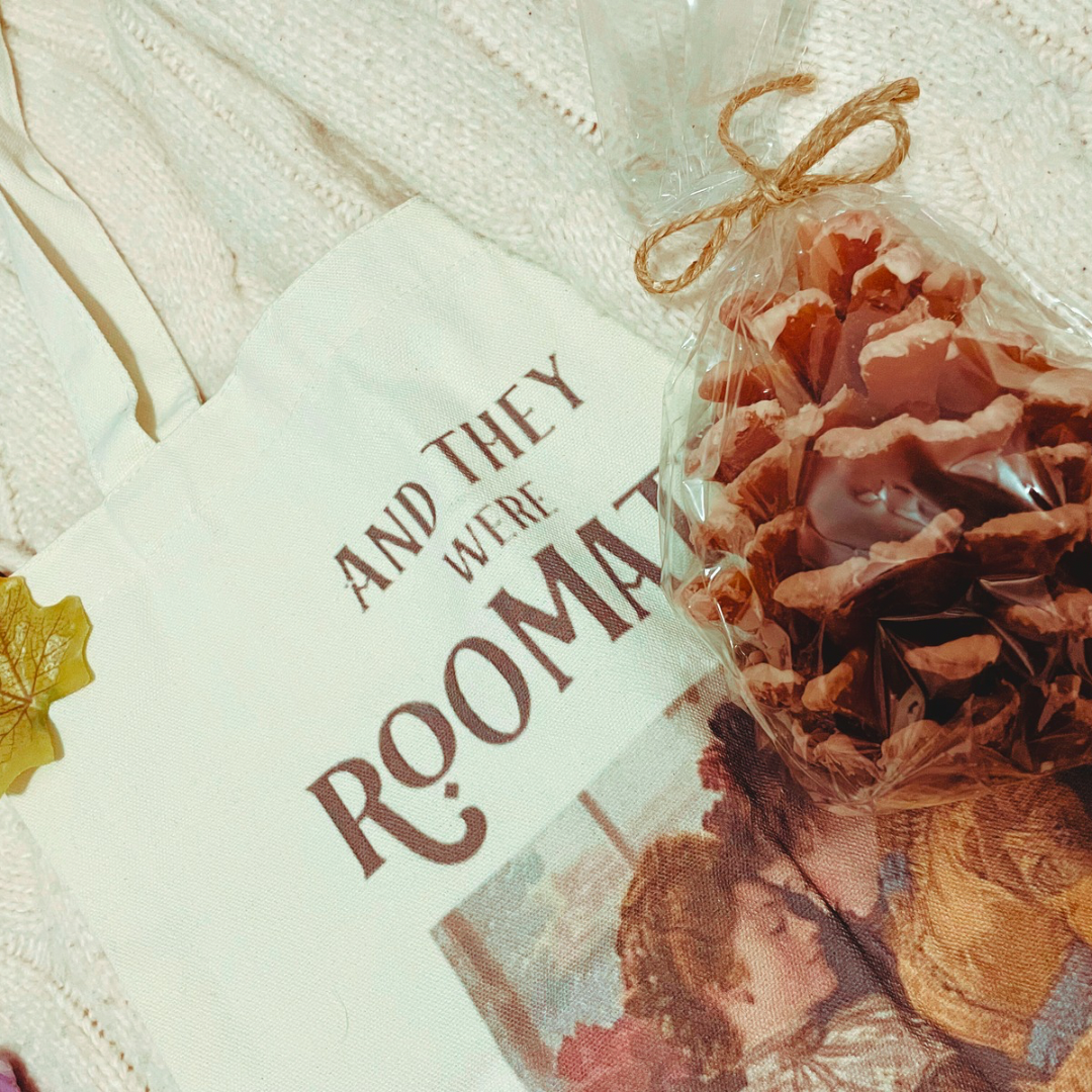 a wrapped pine cone shaped candle lays on an “and they were roommates” tote bag which, in turn, lays on a cream knit cardigan 