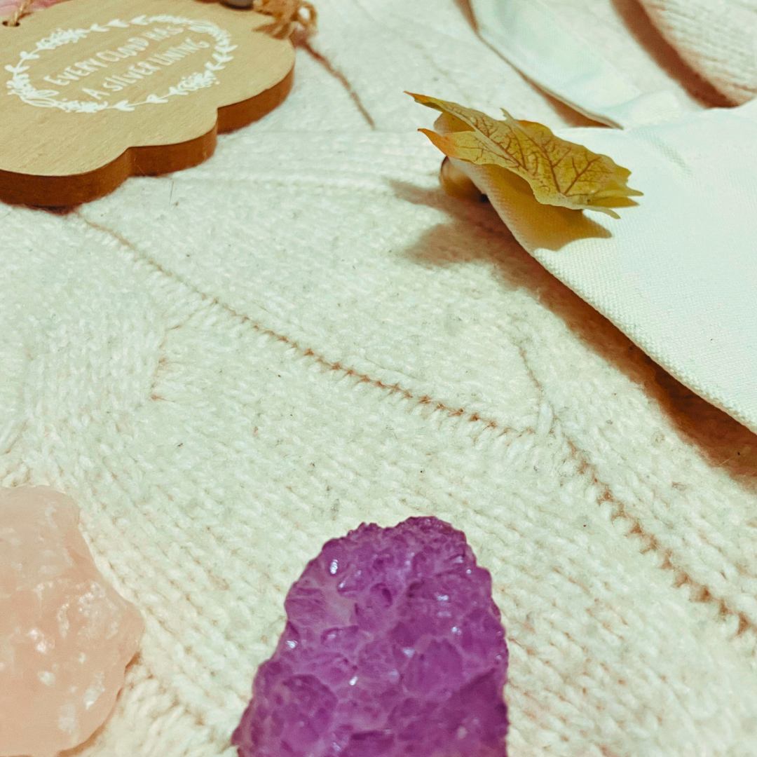 A candle, two crystals, a wooden with “every cloud has a silver lining” and a “and they were roommates” tote bag lay on a cream knit cardigan with some faux leaves