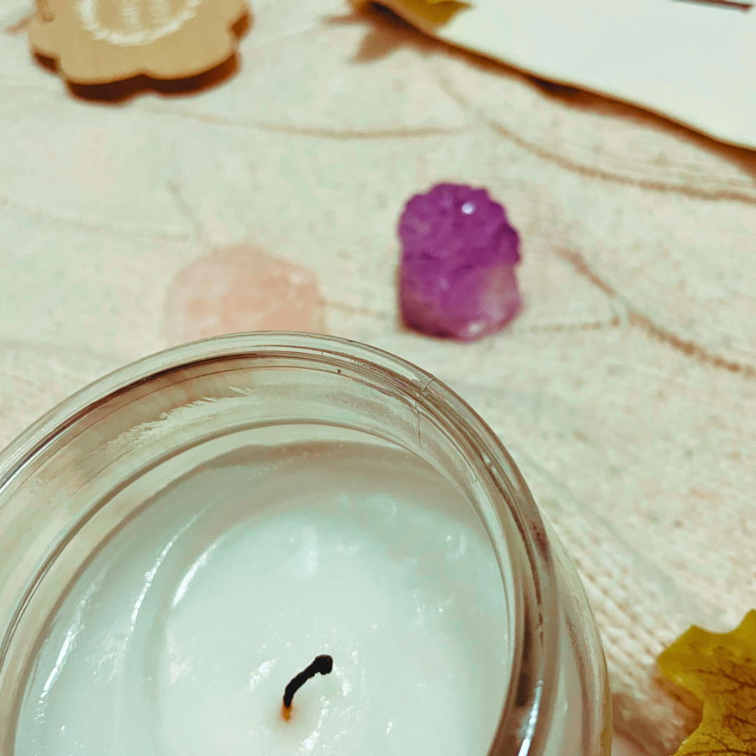 A candle, two crystals, a wooden with “every cloud has a silver lining” and a “and they were roommates” tote bag lay on a cream knit cardigan with some faux leaves