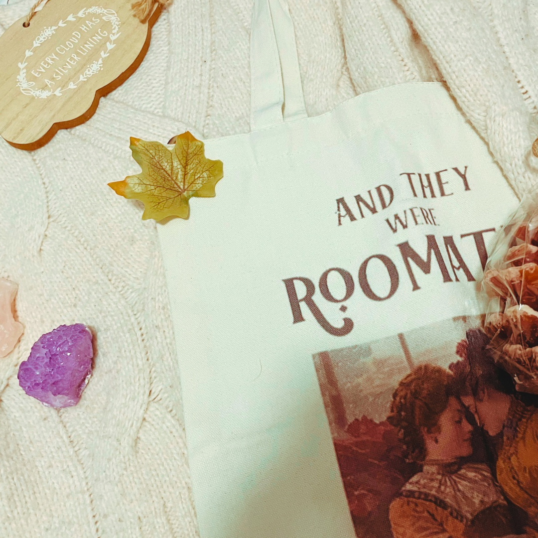 A wrapped pine cone shaped candle, two crystals, a wooden with “every cloud has a silver lining” and a “and they were roommates” tote bag lay on a cream knit cardigan with some faux leaves