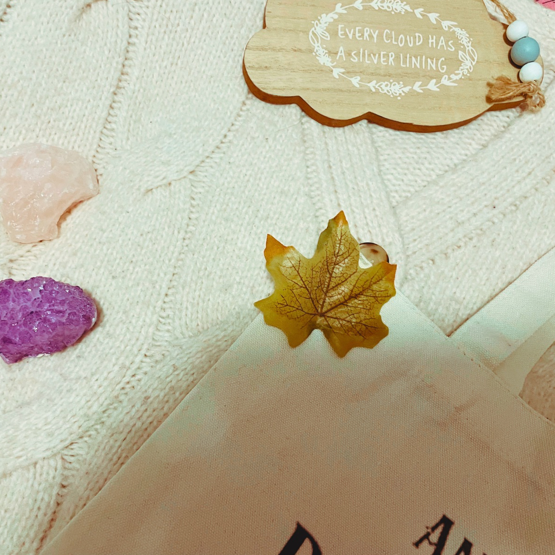 A wrapped pine cone shaped candle, two crystals, a wooden with “every cloud has a silver lining” and a “and they were roommates” tote bag lay on a cream knit cardigan with some faux leaves