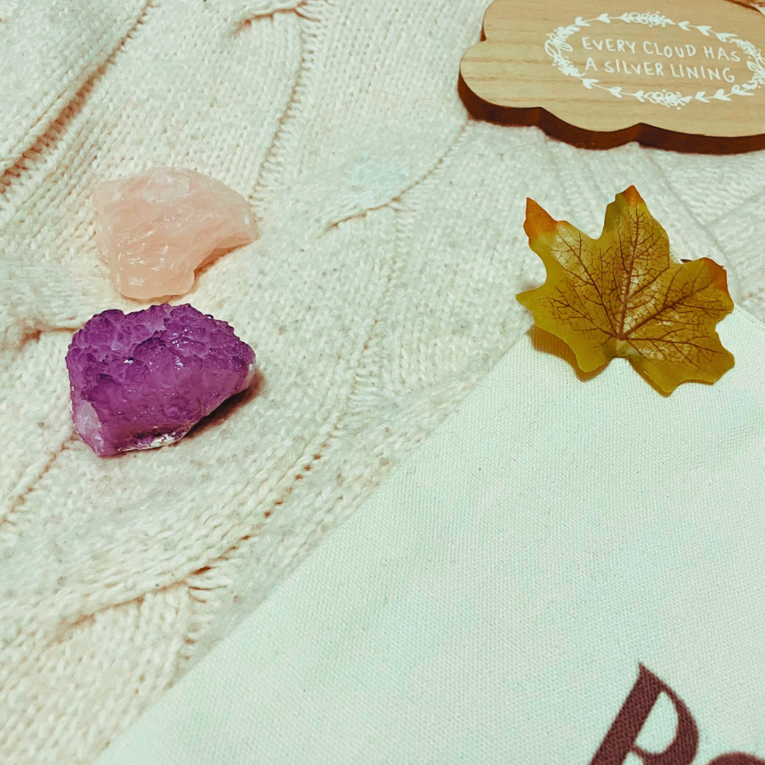 A wrapped pine cone shaped candle, two crystals, a wooden with “every cloud has a silver lining” and a “and they were roommates” tote bag lay on a cream knit cardigan with some faux leaves