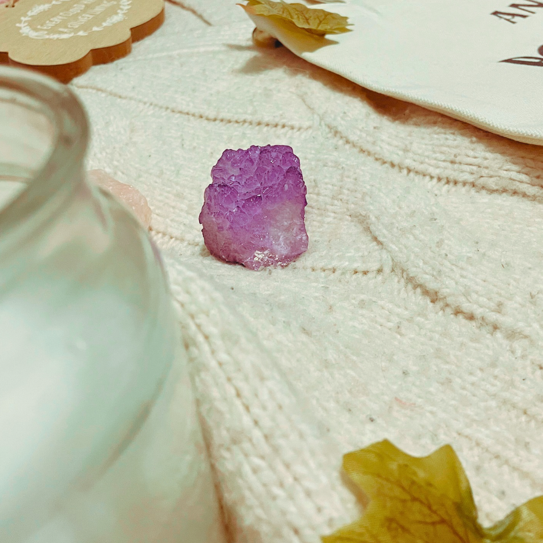 A candle, two crystals, a wooden with “every cloud has a silver lining” and a “and they were roommates” tote bag lay on a cream knit cardigan with some faux leaves
