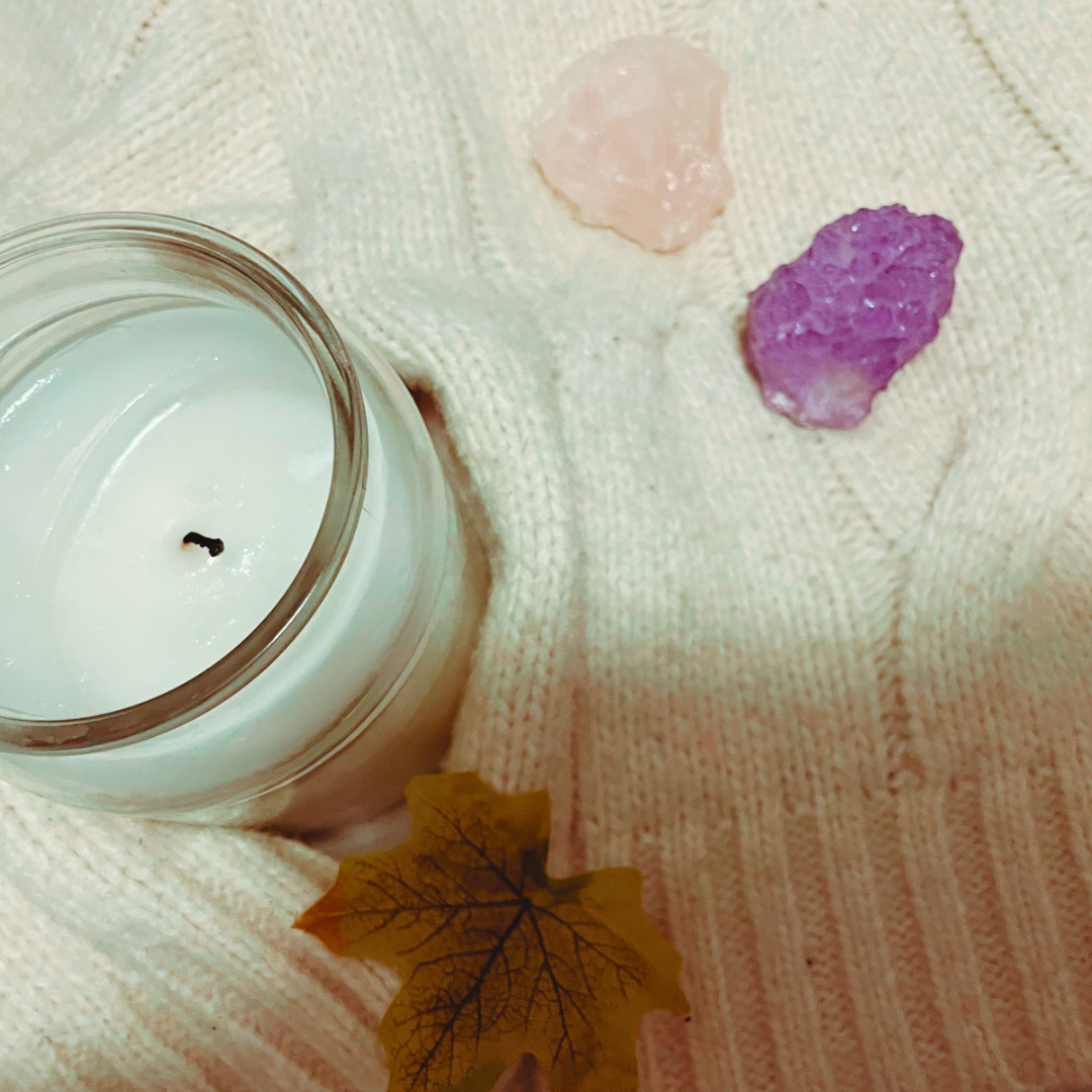 A candle, two crystals, a wooden with “every cloud has a silver lining” and a “and they were roommates” tote bag lay on a cream knit cardigan with some faux leaves