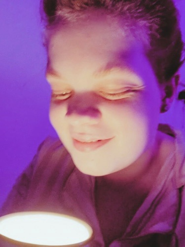 Tig holds a mug candle that they look down at with a big smile 