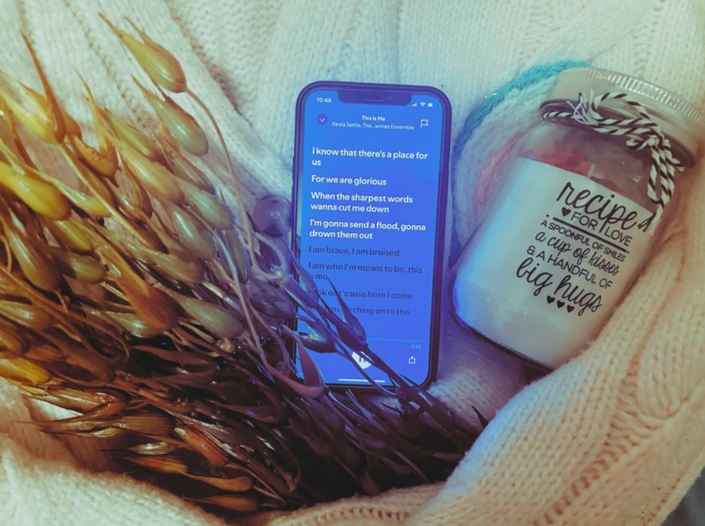 A phone playing “This Is Me”, a faux seaweed plant, a french knitted candle rug and a “Recipe For Love” candle sit within the arms of a cream knitted cardigan.