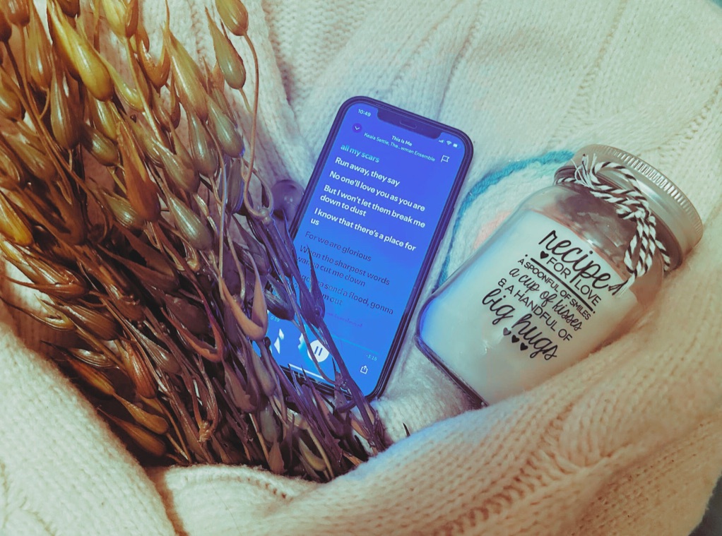 A phone playing “This Is Me”, a faux seaweed plant, a french knitted candle rug and a “Recipe For Love” candle sit within the arms of a cream knitted cardigan.