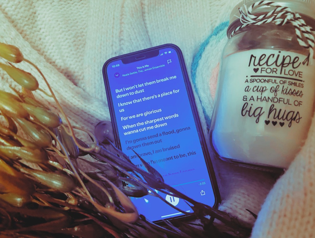 A phone playing “This Is Me”, a faux seaweed plant, a french knitted candle rug and a “Recipe For Love” candle sit within the arms of a cream knitted cardigan.
