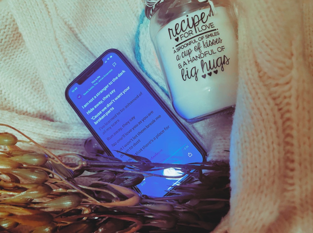 A phone playing “This Is Me”, a faux seaweed plant, a french knitted candle rug and a “Recipe For Love” candle sit within the arms of a cream knitted cardigan.